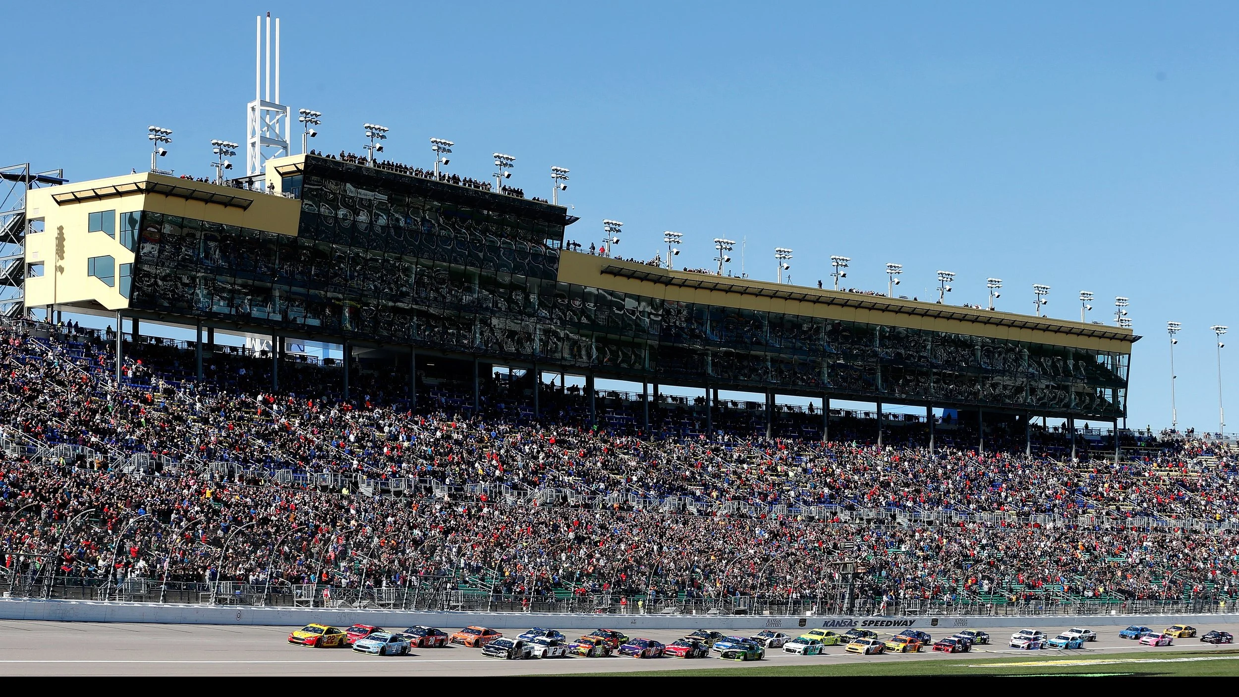 Race cars on track at Kansas Speedway with packed grandstands and a large press box under a clear blue sky.