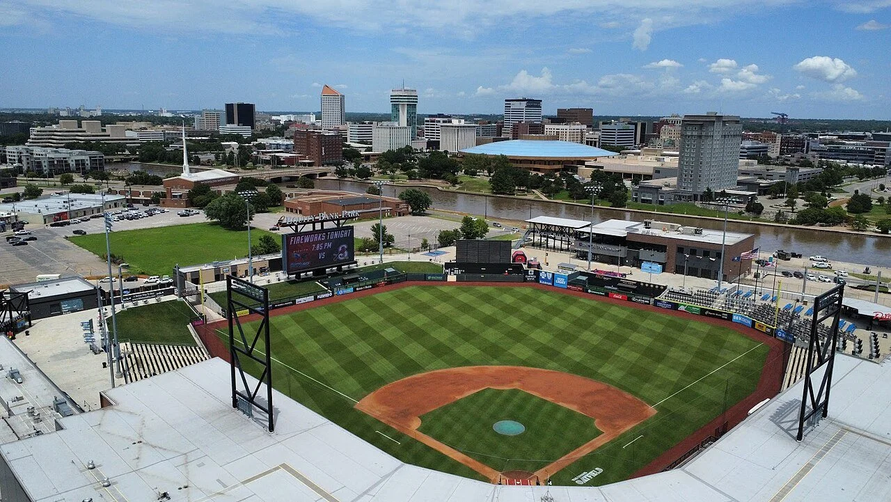 An aerial view of a baseball stadium in downtown, with city buildings and a waterway in the background.