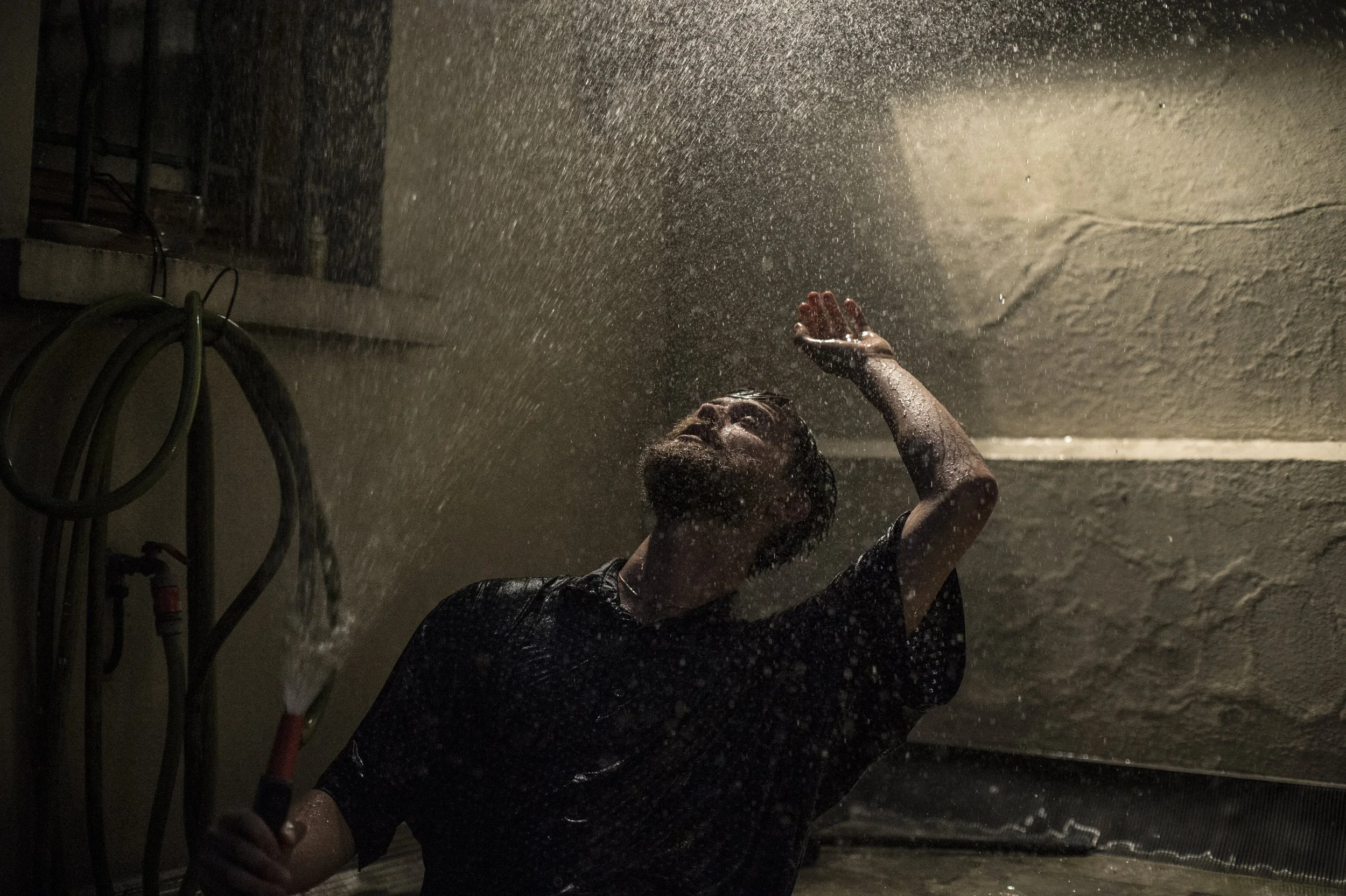 A man standing in the rain, tilting his head back with one arm raised to shield his face, holding a garden hose in his other hand.