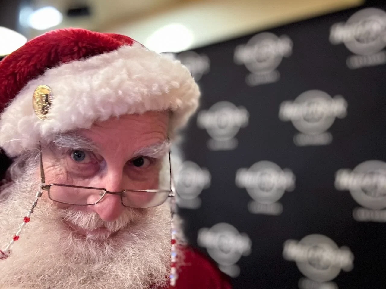 Close-up of a man dressed as Santa Claus, wearing glasses and a red Santa hat with white fur trim, standing in front of a black backdrop with logos.
