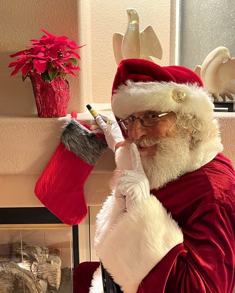 A man dressed as Santa Claus, smiling and looking mischievous, tuning a Christmas stocking hanging on the mantel, decorated with a red poinsettia plant and ceramic dove sculptures in the background.