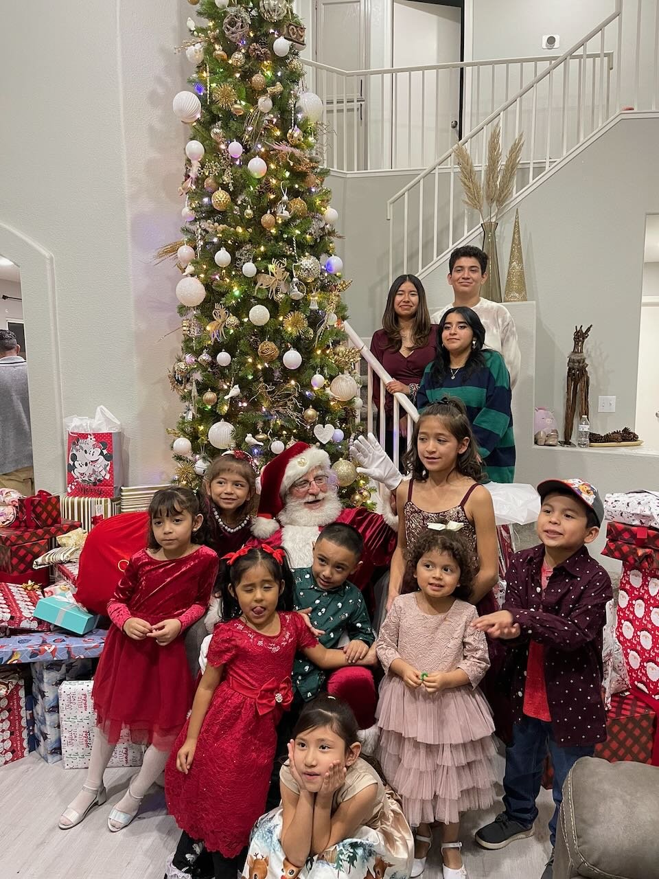 Group of children and two adults with Santa Claus around a decorated Christmas tree, with wrapped presents underneath, on a festive holiday evening.