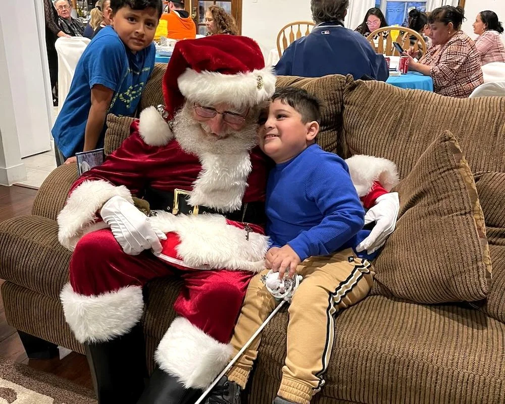 Two young boys sitting on a brown couch, hugging Santa Claus who is dressed in a traditional red and white suit. A boy in a blue sweater is smiling while Santa, with glasses and a white beard, is leaning in for a photo. In the background, people are seated at tables in a busy indoor setting.