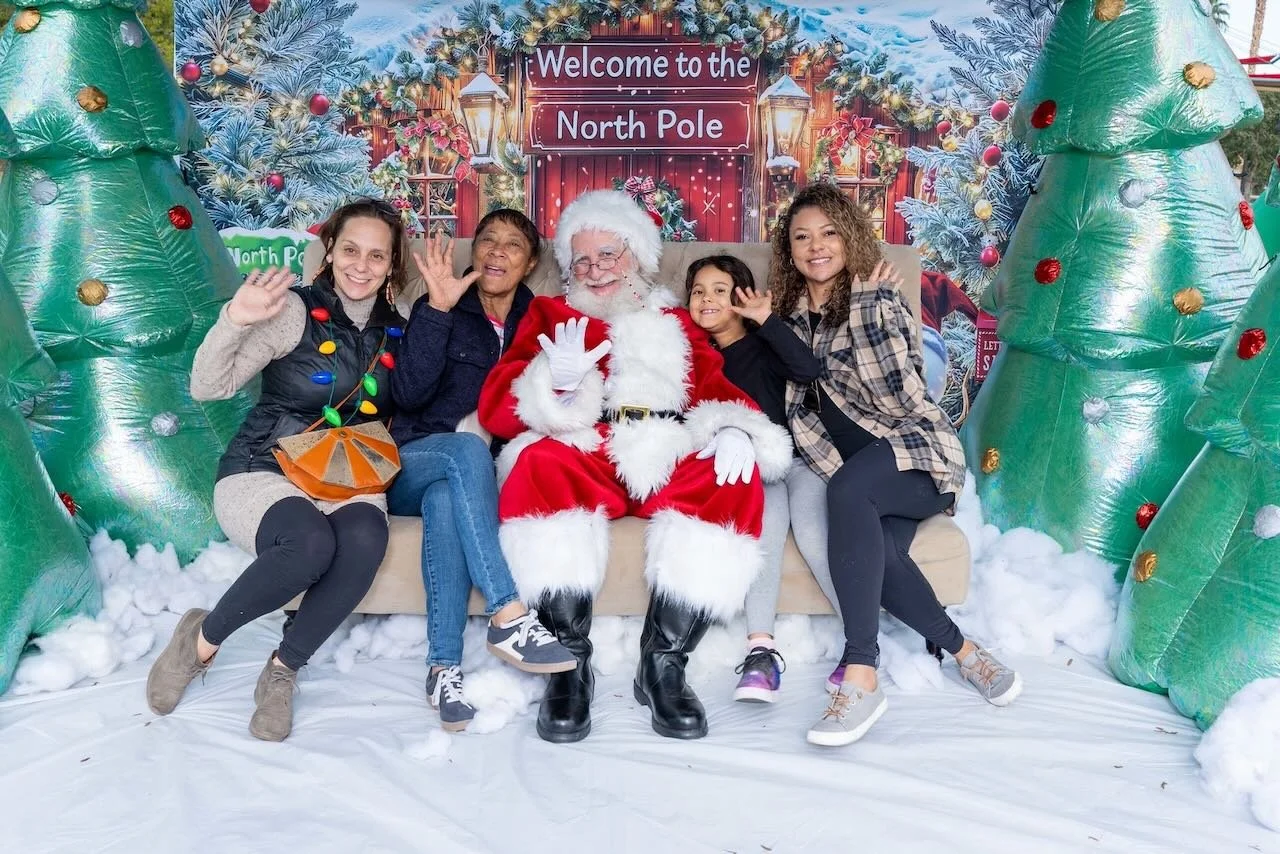 Four women and Santa Claus sitting on a bench, waving, with Christmas decorations and a sign that reads "Welcome to the North Pole" behind them.