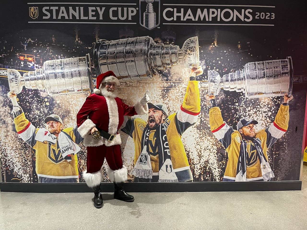 Santa Claus dressed in a red suit and black boots standing in front of a large banner that reads 'Stanley Cup Champions 2023'. The banner features three hockey players in yellow and black jerseys celebrating with the Stanley Cup trophy amid fireworks and confetti. One player is holding the trophy overhead, another is holding it to the side, and the third is in the middle celebrating loudly.