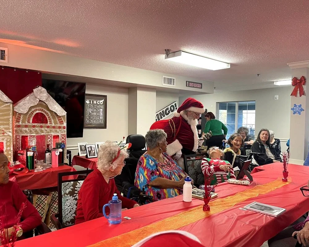 A Christmas gathering with elderly people seated around a table decorated with red and gold tablecloth and centerpieces, a man dressed as Santa Claus standing, a gingerbread house decoration on the wall, and Christmas decorations like bows and snowflakes in the room.