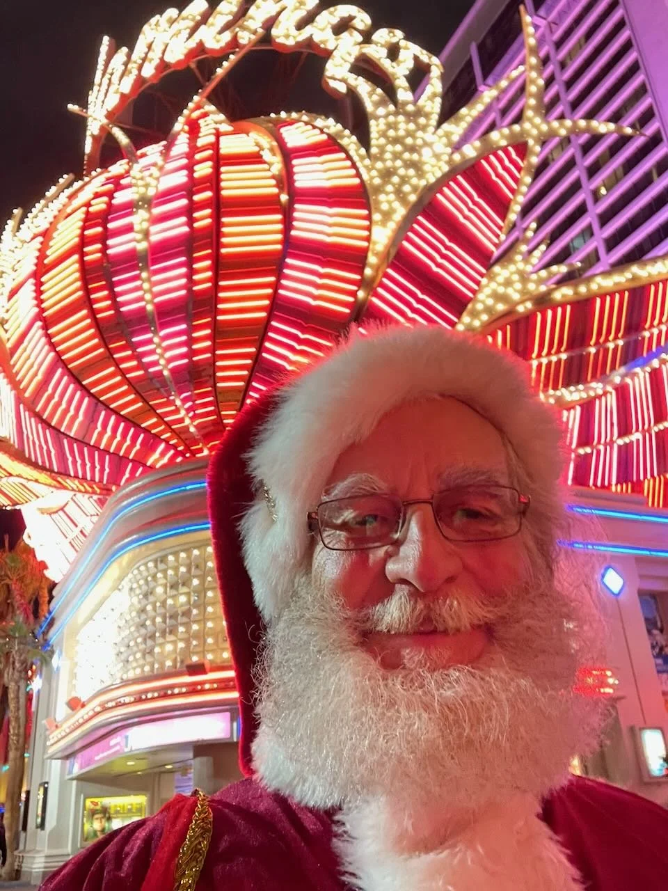 A man dressed as Santa Claus smiling at night in front of a brightly lit, colorful carnival or amusement park ride with neon lights.
