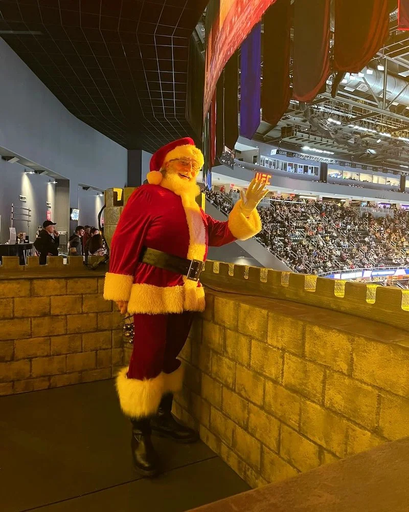 Santa Claus in a red suit with white fur trim, waving at an event with a large crowd and banners overhead.