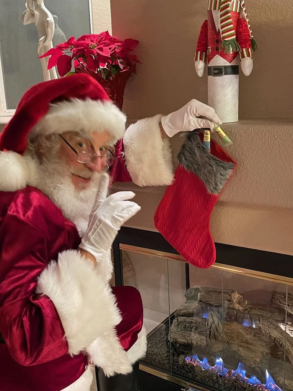 Santa Claus in a red velvet outfit with white fur trim, wearing glasses and gloves, smiling and pointing to a red Christmas stocking hanging above a fireplace, with a poinsettia plant and elf decoration in the background.