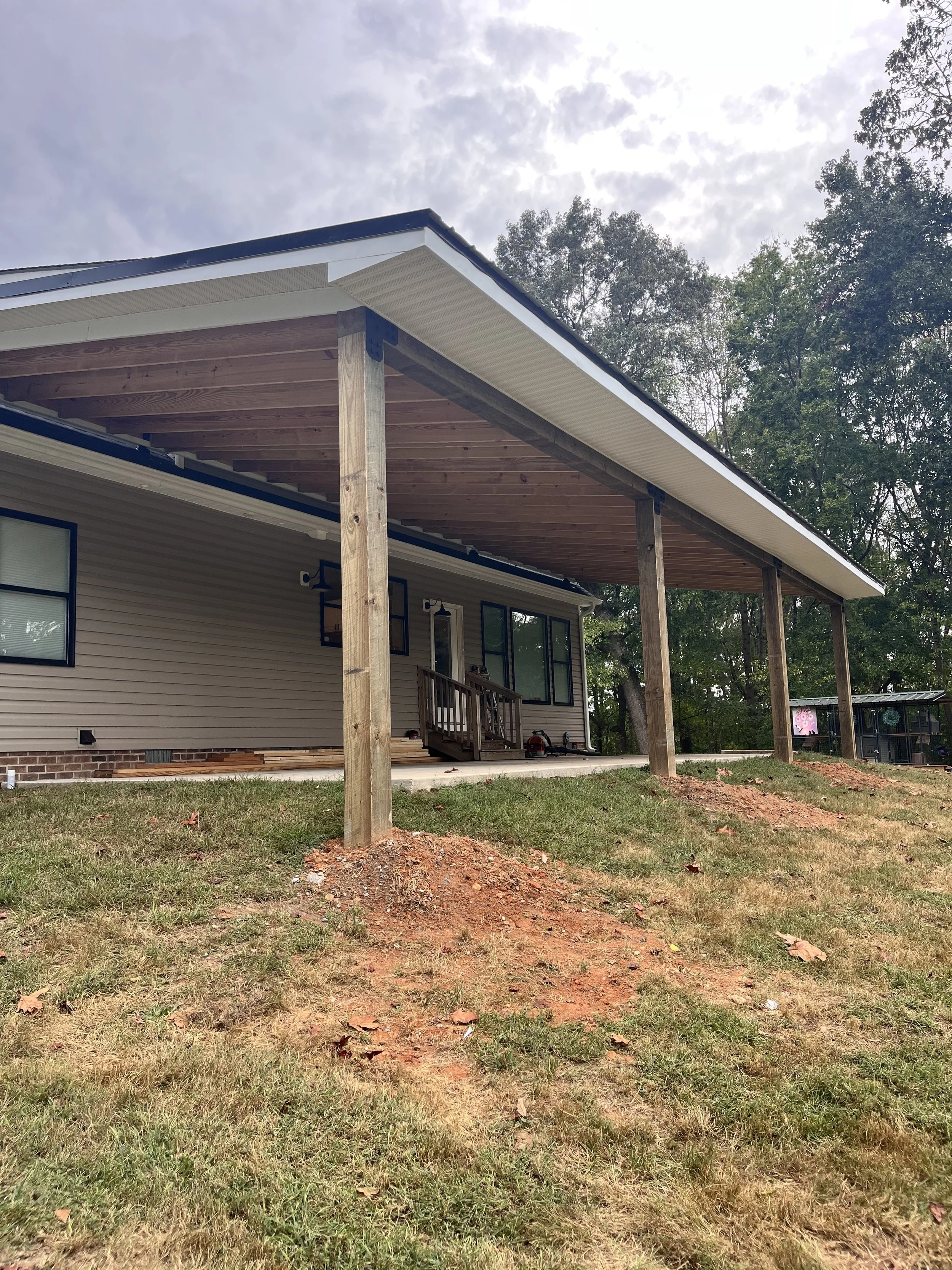 Newly constructed house with a covered porch supported by wooden posts, with some construction materials on the ground. The house has beige siding, black-trimmed windows, and a nearby shed, with trees and a cloudy sky in the background.
