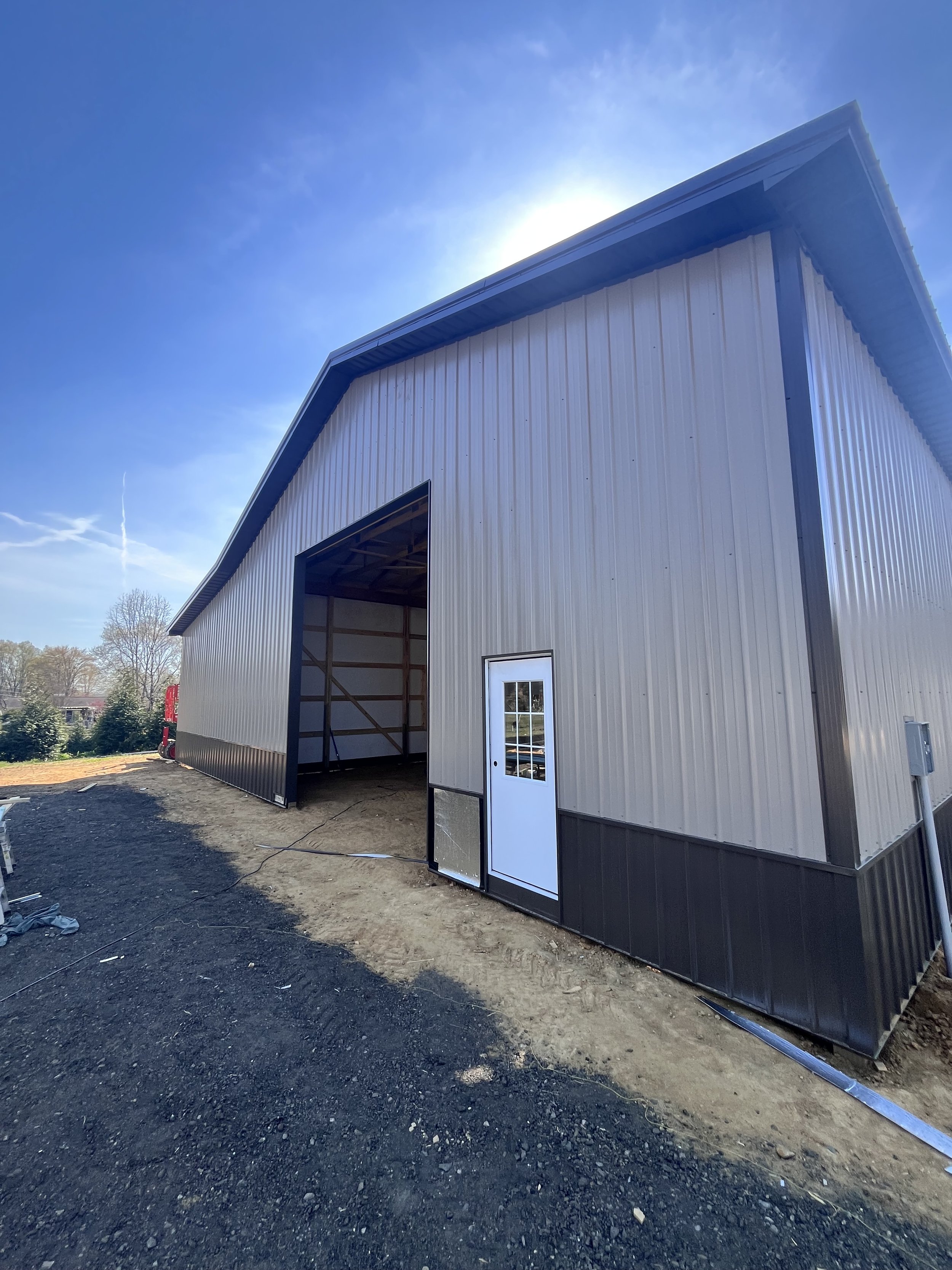 Under construction metal building with an open garage door and a side door, surrounded by dirt and gravel, with trees and a blue sky in the background.