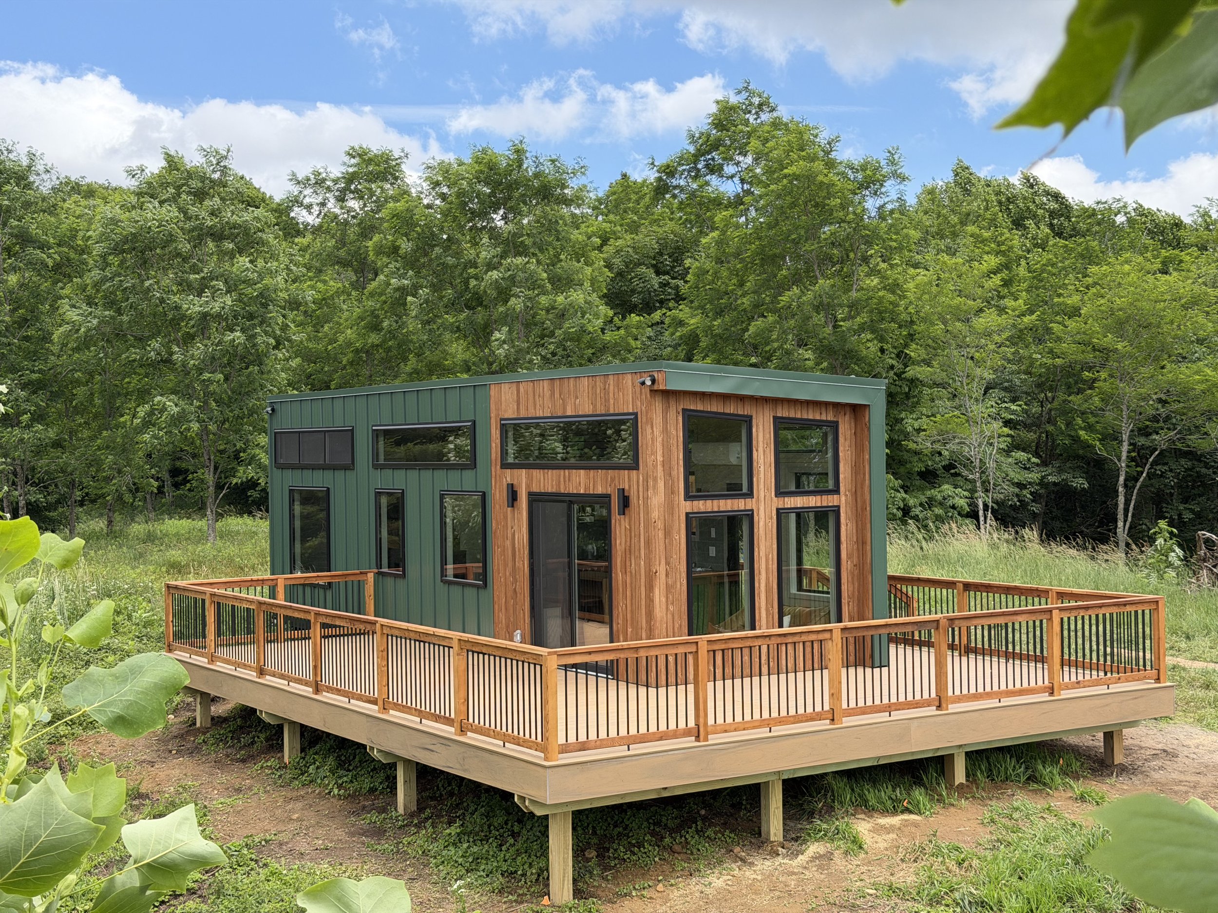 Modern tiny house with green metal siding and wooden accents, open porch with wooden railing, situated among trees and greenery under a partly cloudy sky.