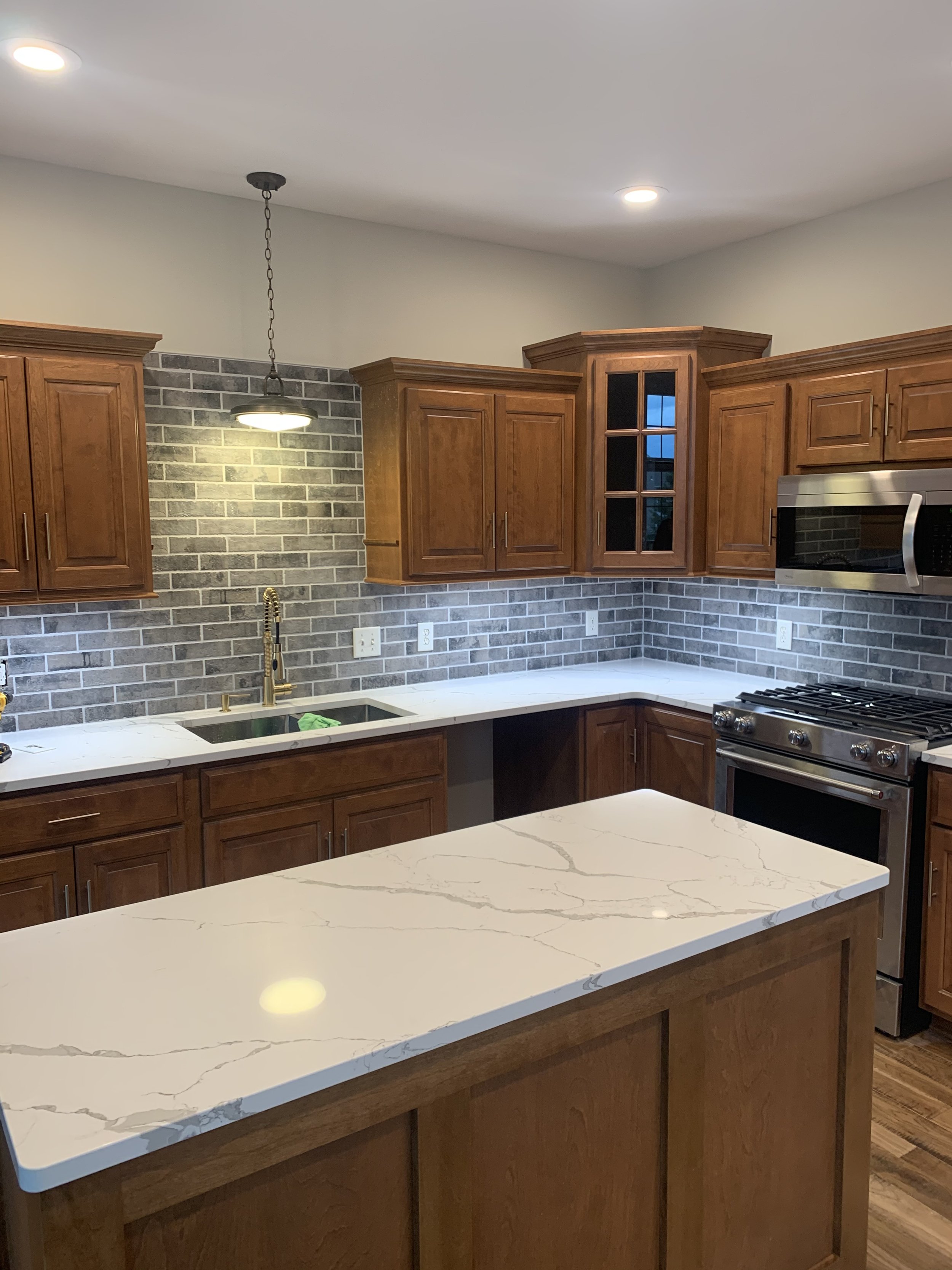 A modern kitchen with wooden cabinets, a white marble countertop, a stainless steel microwave, a gas stove, a stovetop faucet, and a gray brick backsplash. There is a hanging light fixture above the sink area and recessed ceiling lights.