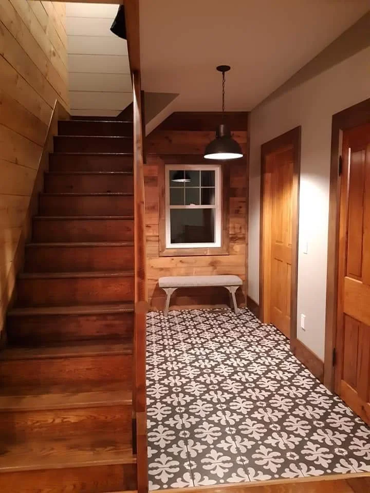 Interior view of a house entryway with wooden stairs on the left, a small window, a bench, and a pendant light hanging from the ceiling, with decorative patterned tile flooring.