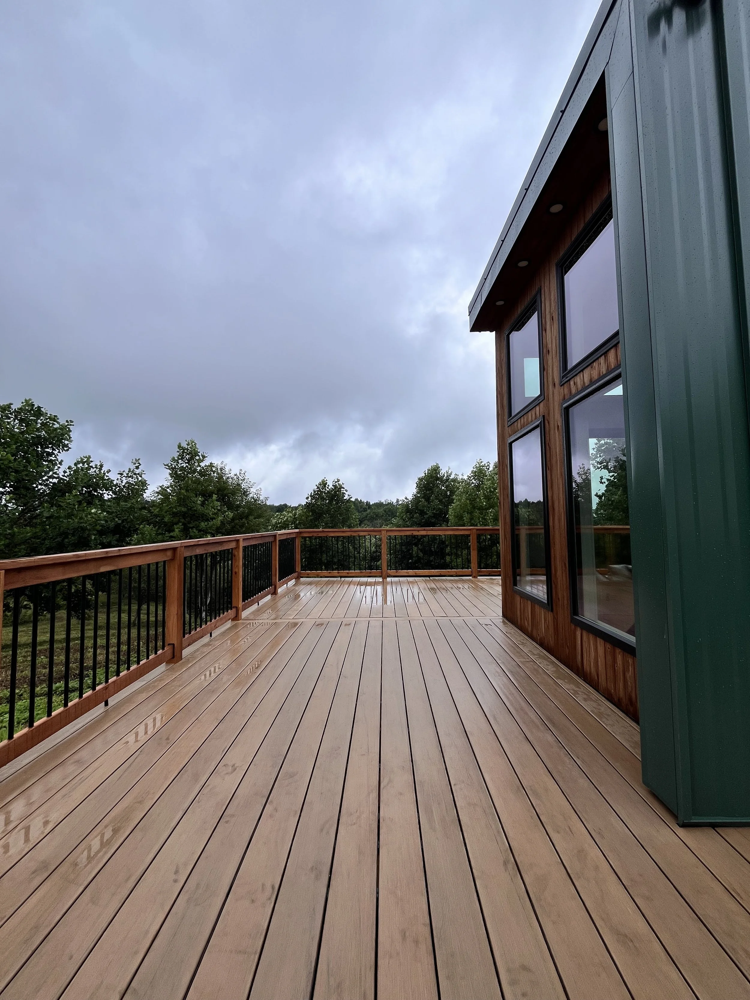 A modern house with large windows, a green exterior wall, and a wooden deck with a railing, overlooking a green, tree-filled landscape under a cloudy sky.