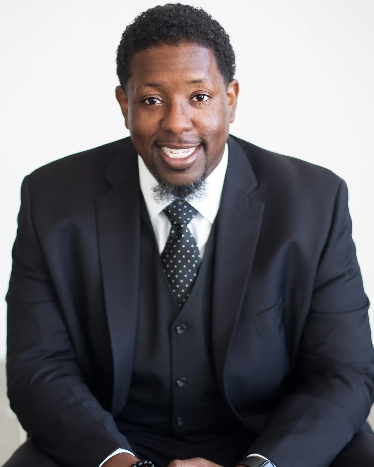 A man in a black suit, white shirt, and black polka dot tie sitting and smiling at the camera.