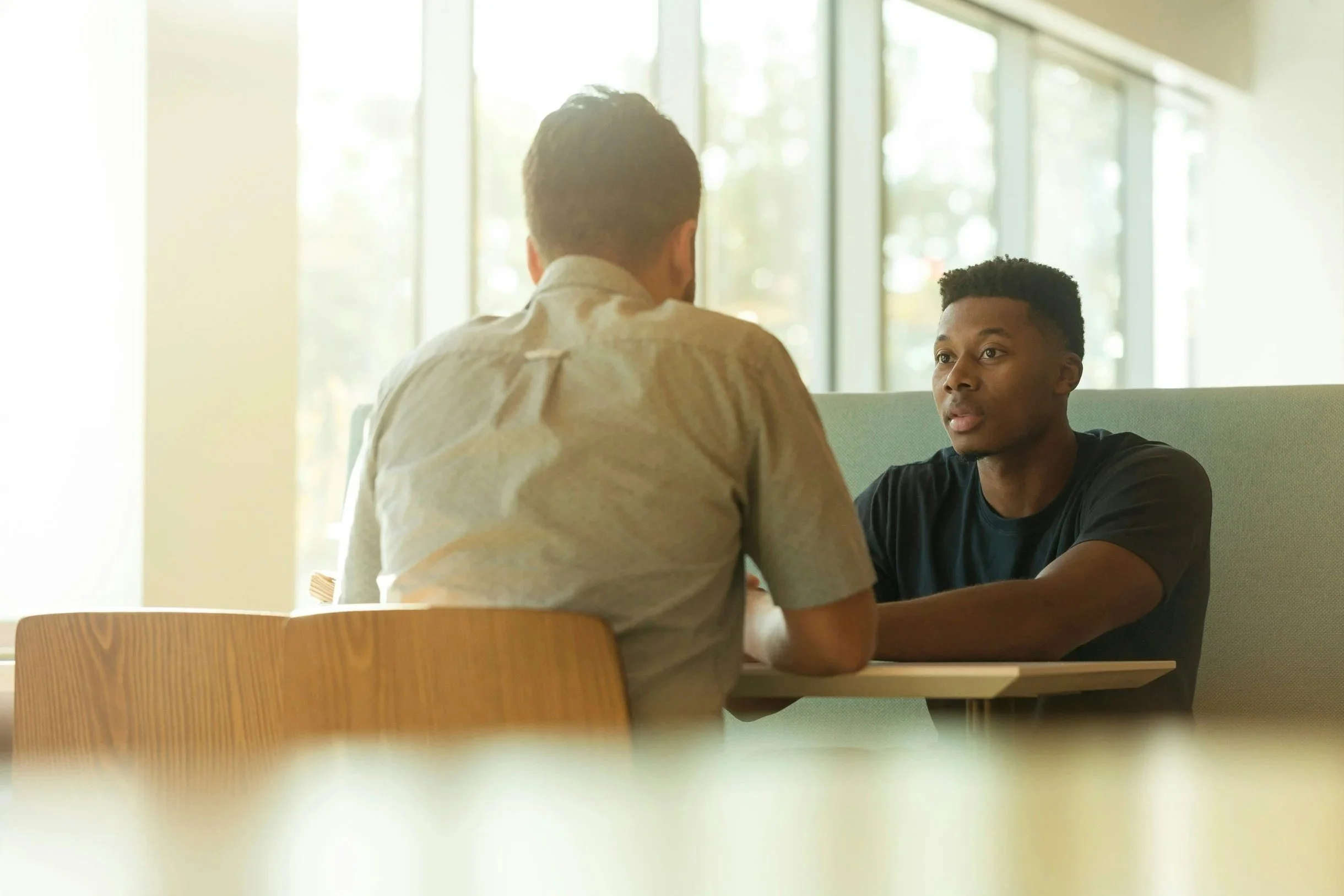 Two men having a serious conversation in a bright indoor setting with large windows, one facing the camera and the other with back to camera.