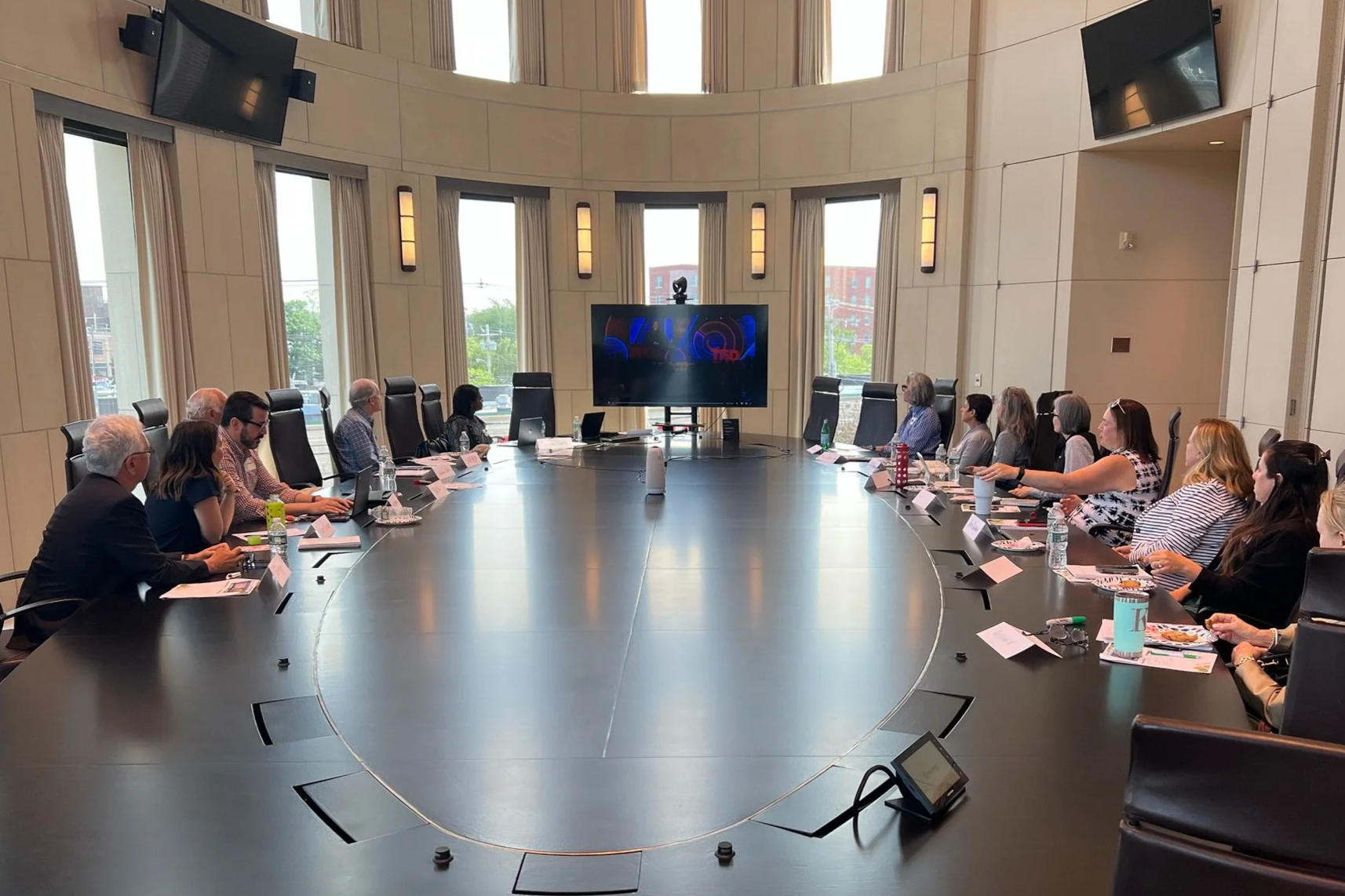 A group of people seated around a large oval conference table in a bright, modern meeting room with large windows, a big TV screen, and wall-mounted monitors.