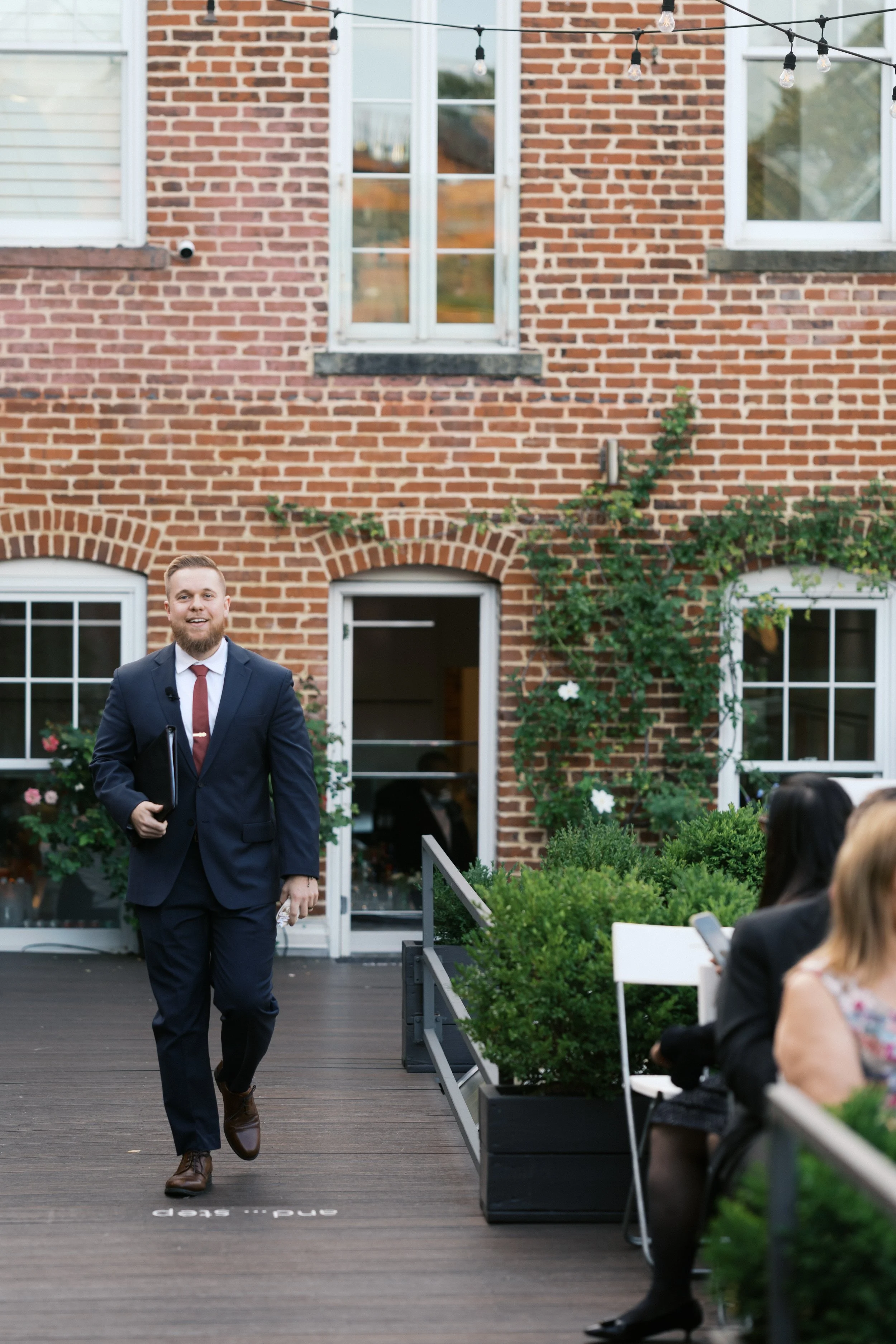 A man in a navy suit and red tie walking on a wooden deck outside a brick building, holding a folder, with people seated nearby.