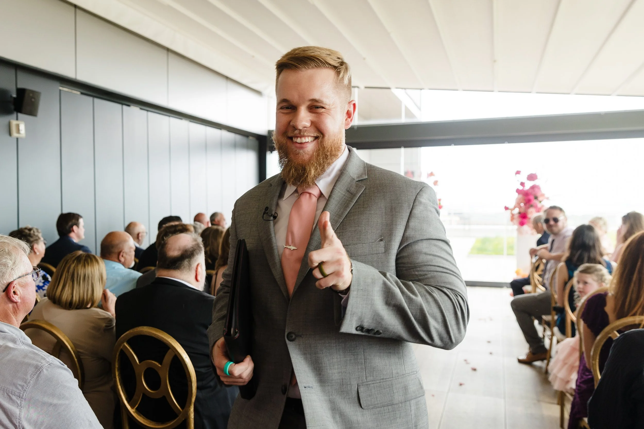 A smiling man with a beard in a gray suit points towards the camera at a formal event with seated guests in the background.