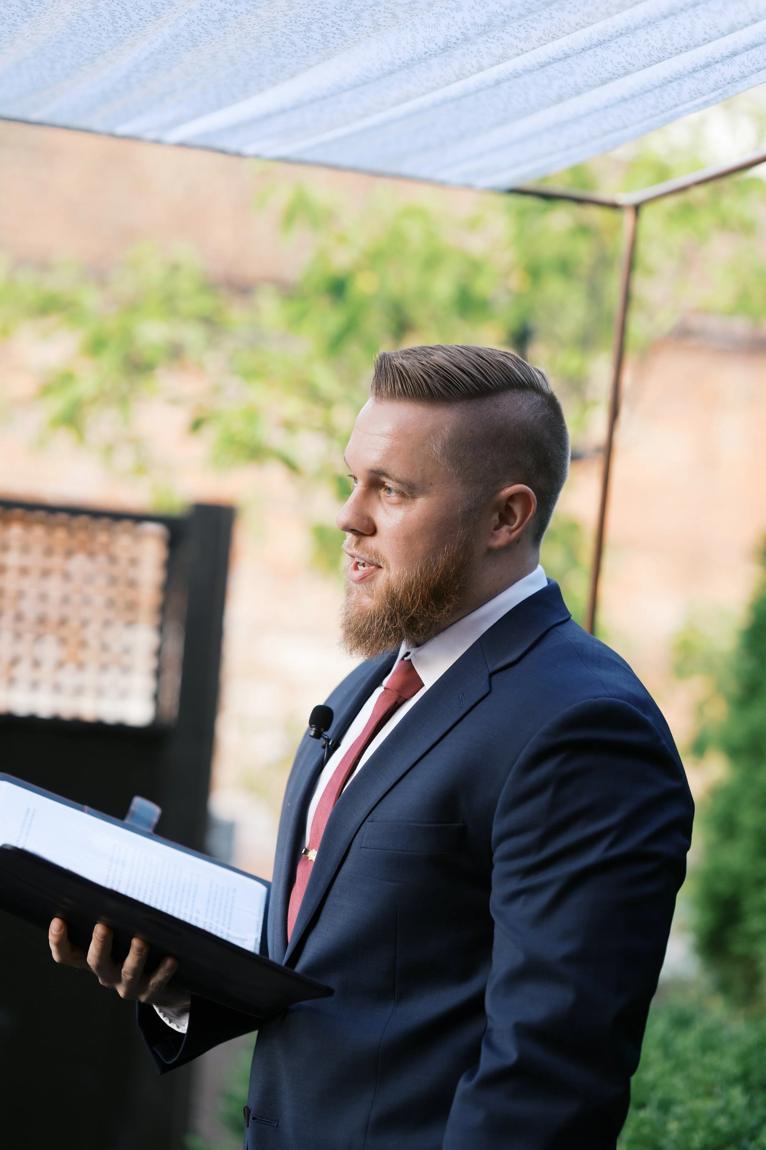 A man giving a speech outdoors, holding a binder with papers, dressed in a navy suit and pink tie, with a beard and styled hair, standing under a tent with green trees in the background.