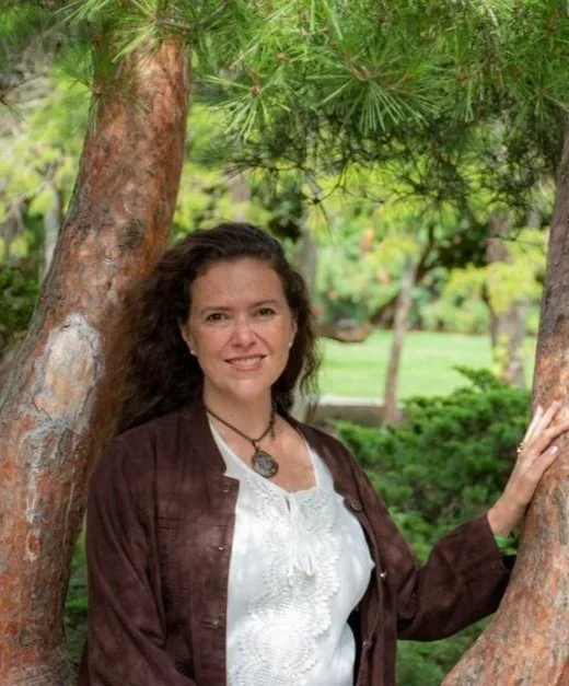 Woman with curly hair standing outdoors between two trees, smiling at the camera, wearing a brown jacket and white top, with a necklace, in a lush green park.