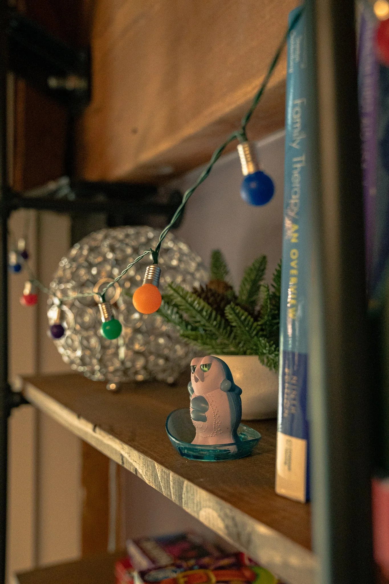 A small figurine of a pink bird with glasses sitting in a blue dish on a wooden shelf. Behind it, there is a small green plant, a crystal-like spherical lamp, a string of colorful lights, and some books.