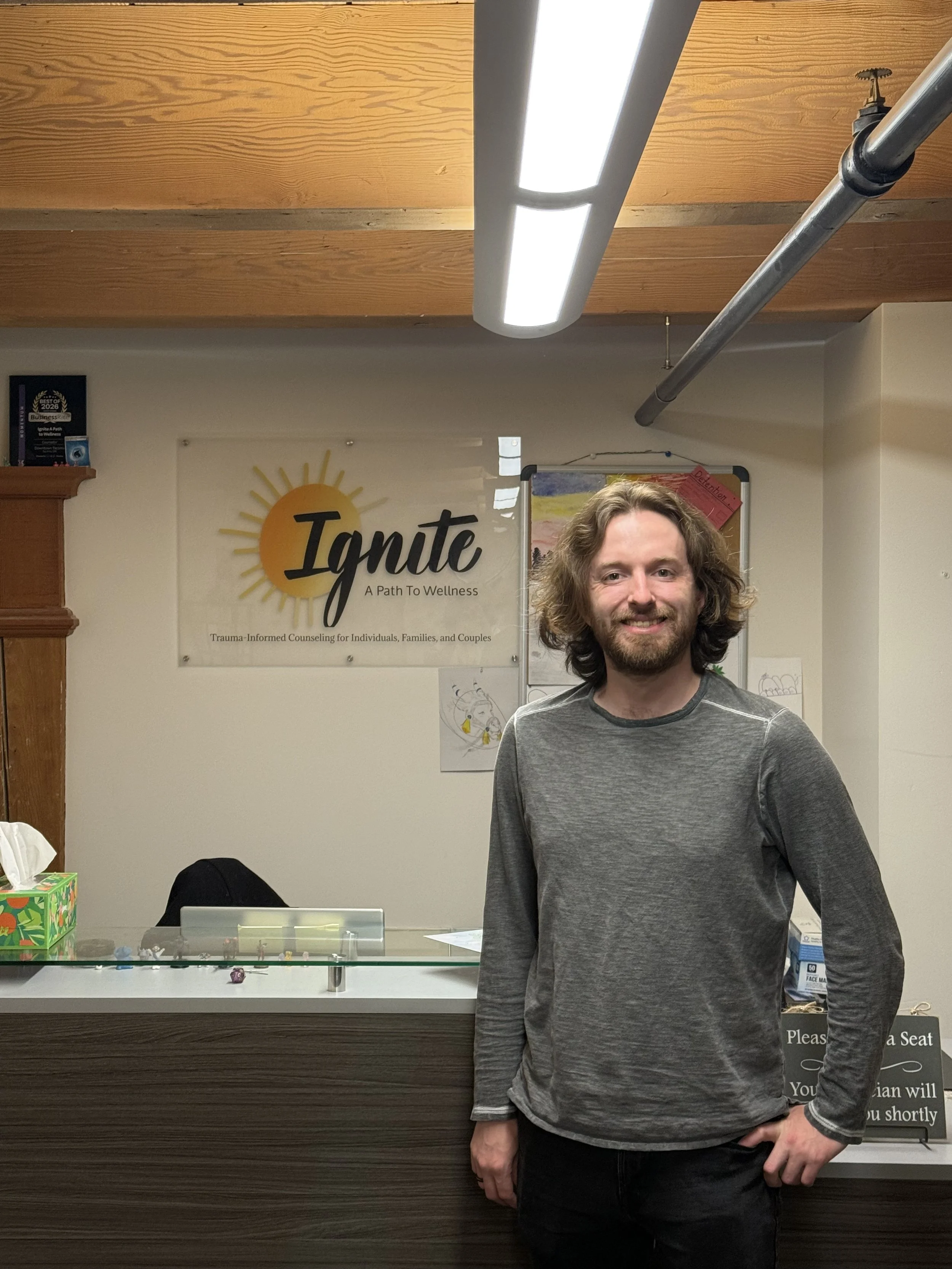 A man with long, curly brown hair and a beard, wearing a gray long-sleeve shirt, standing in front of a reception desk with a sign that reads 'Ignite: A Path to Wellness' and a poster on the wall behind him.