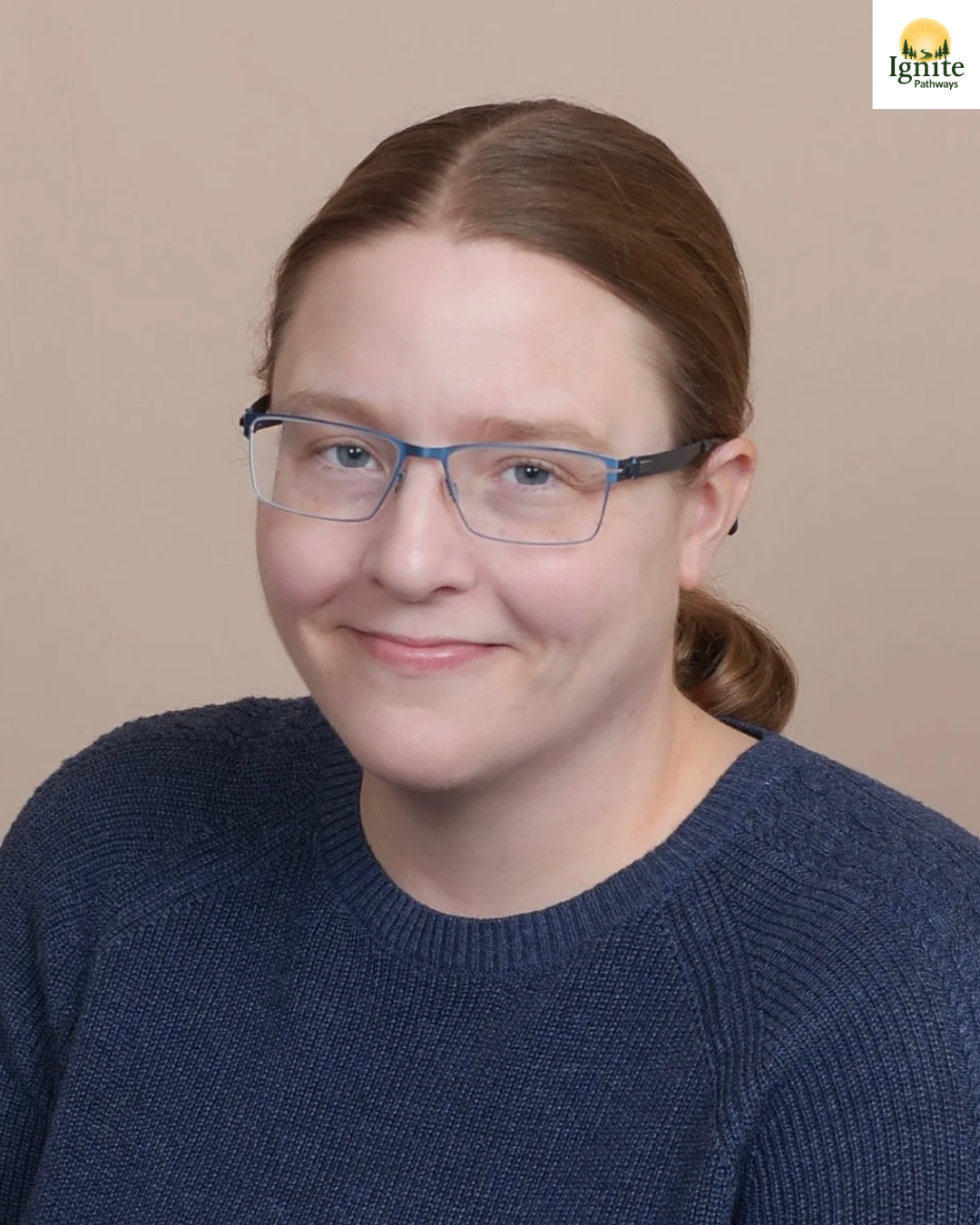 Headshot of a woman with light skin, red hair tied back, wearing glasses and a dark blue sweater, smiling against a beige background. The Ignite Pathways logo is in the top right corner.