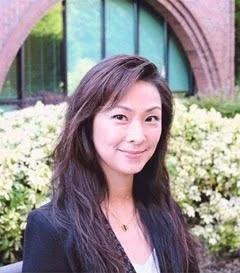 A woman with long dark hair smiling outdoors in front of a building with large windows and white flowers.