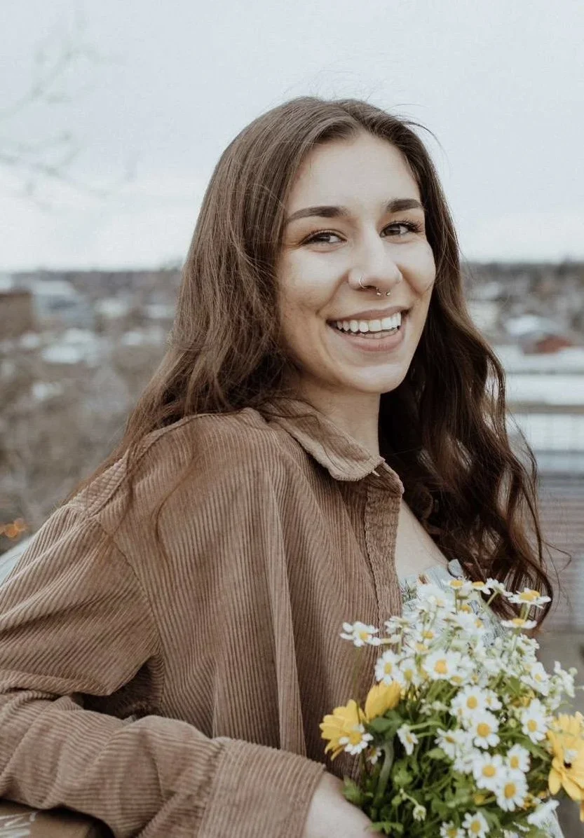 A young woman with long brown hair and a nose ring, smiling and holding a bouquet of white daisies and yellow flowers, outdoors with a blurred cityscape background.