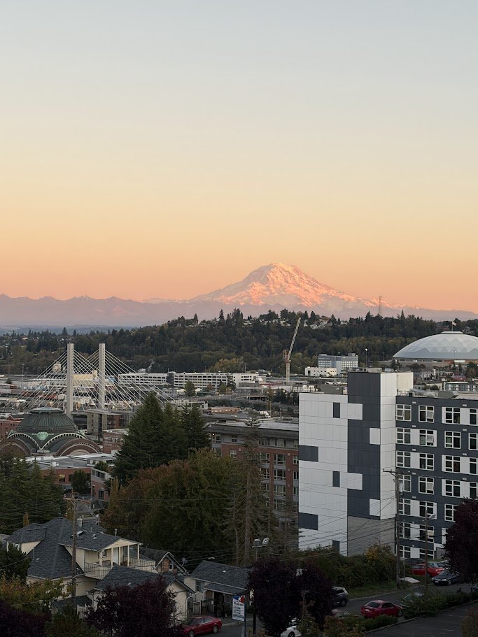 Cityscape with downtown buildings, trees, and a bridge in the foreground, and snow-capped Mount Rainier in the background at sunset.