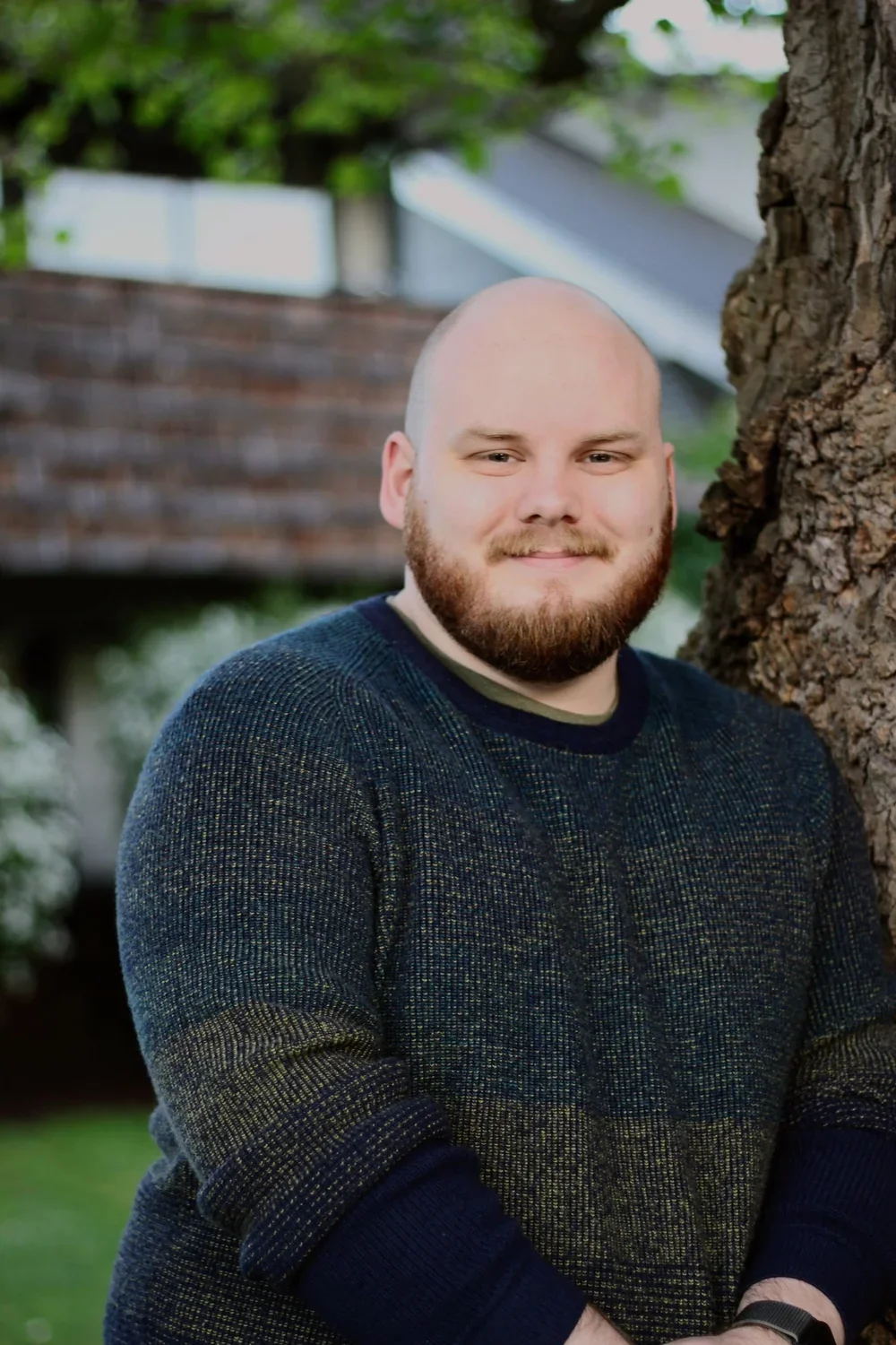 A man with a beard and bald head, wearing a dark sweater, standing outdoors next to a tree, with a blurred background of a brick building and green foliage.