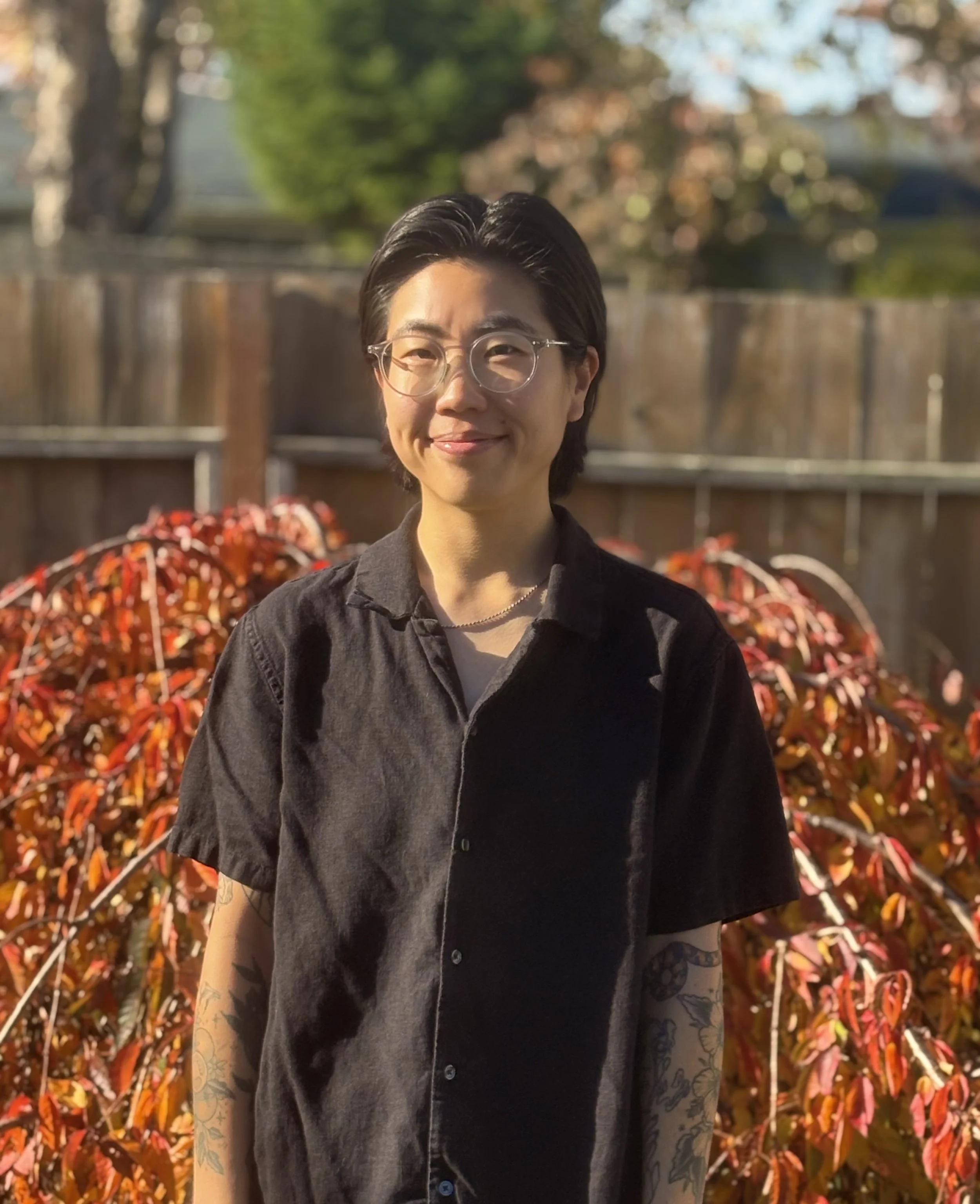 Portrait of a young person with short black hair, wearing round glasses, a black button-up shirt, and visible tattoos on arms, standing outdoors during fall with a wooden fence and colorful autumn leaves in the background.
