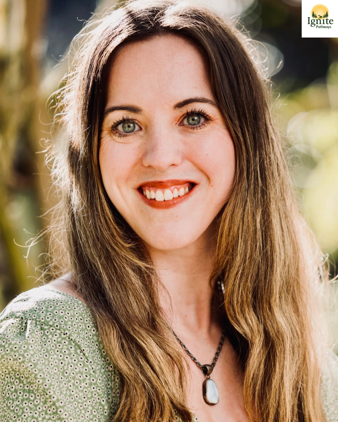 Close-up of a woman with long brown hair, blue eyes, and a nose piercing, smiling outdoors on a sunny day.
