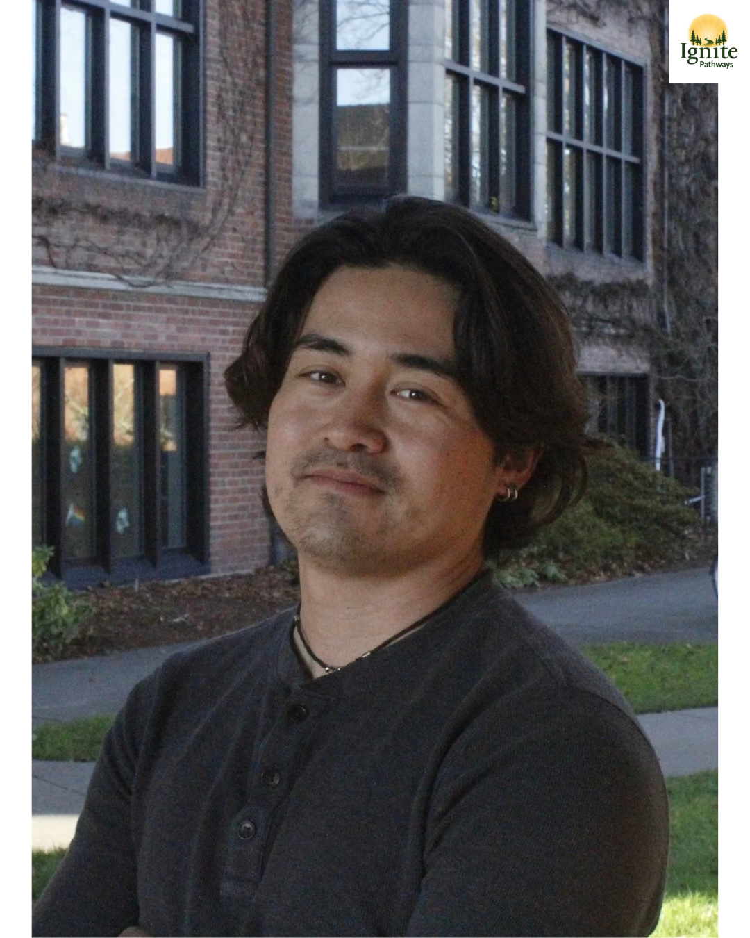 A young man with dark, wavy hair and earrings, standing outdoors in front of a brick building with large windows, during late afternoon or early evening.