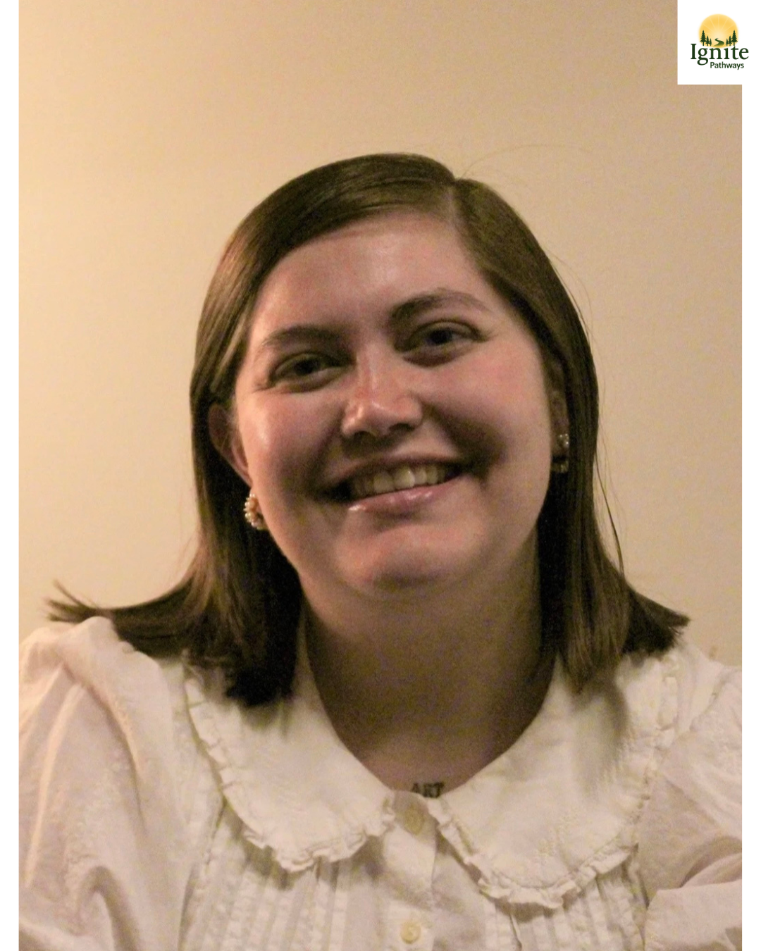 A smiling young woman with shoulder-length brown hair, wearing pearl earrings, a white blouse with a ruffled collar, and a small necklace. The background is plain and beige. The Ignite Pathways logo is in the top right corner.