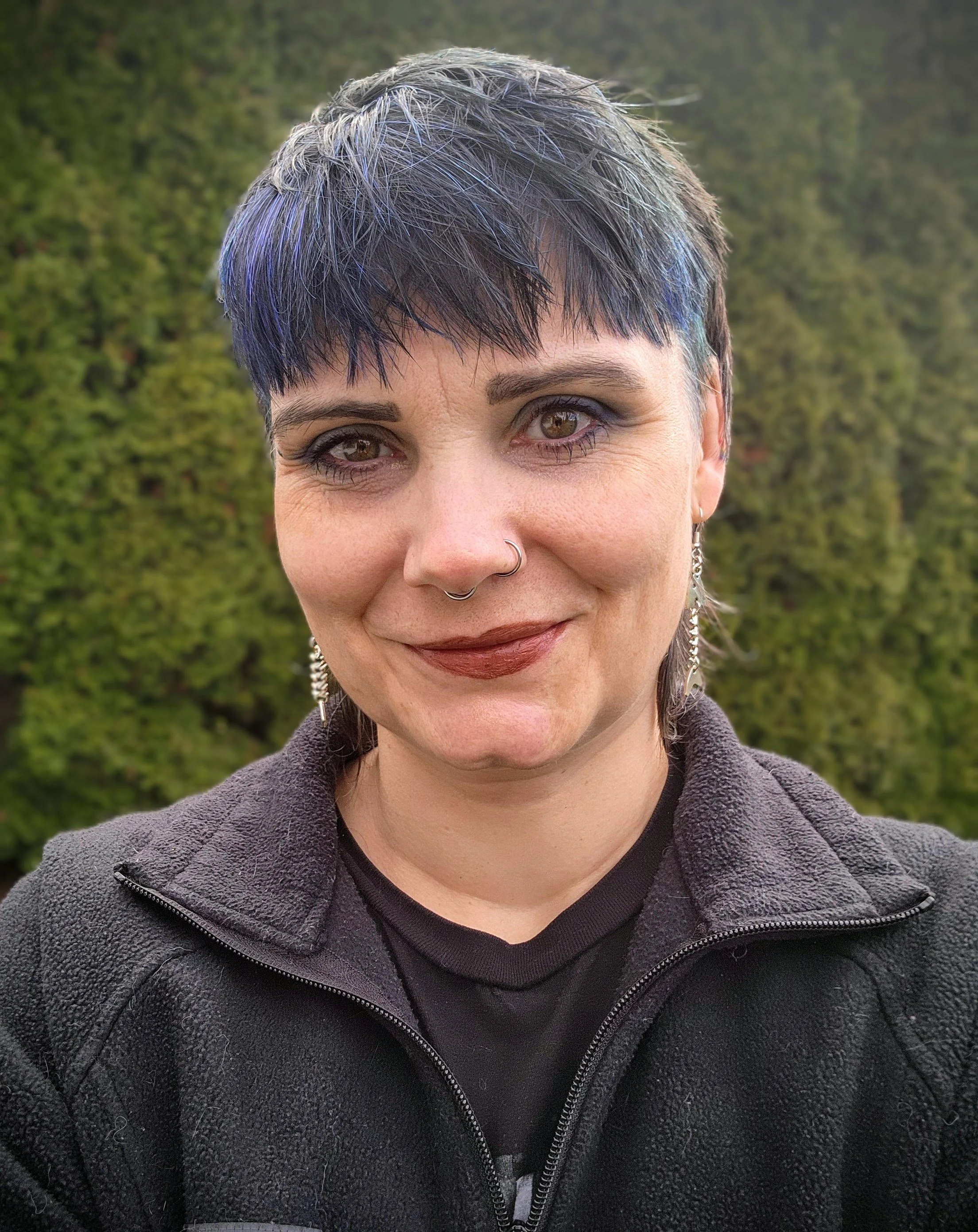 Close-up of a woman's face with short, dark hair with streaks of purple, brown eyes, wearing earrings and a nose ring, smiling slightly, outdoors with a green forest background.