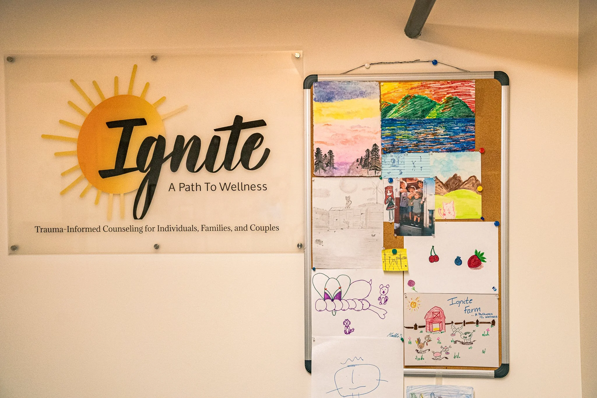 Whiteboard with colorful landscape and nature drawings, photos of children, and farm-themed artwork, alongside a sign reading 'Ignite: A Path To Wellness' for trauma-informed counseling.