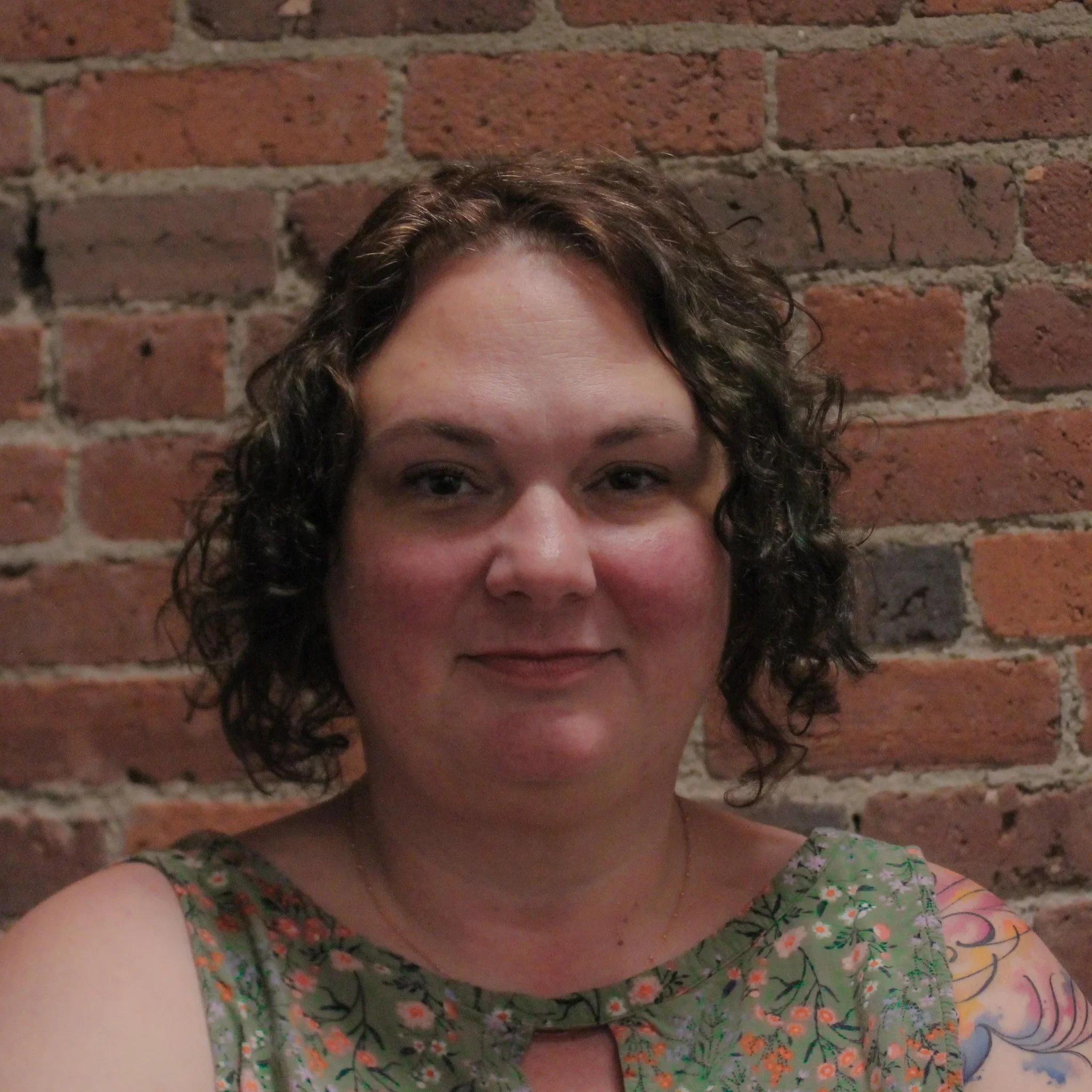 A woman with curly brown hair wearing a floral dress standing in front of a brick wall.