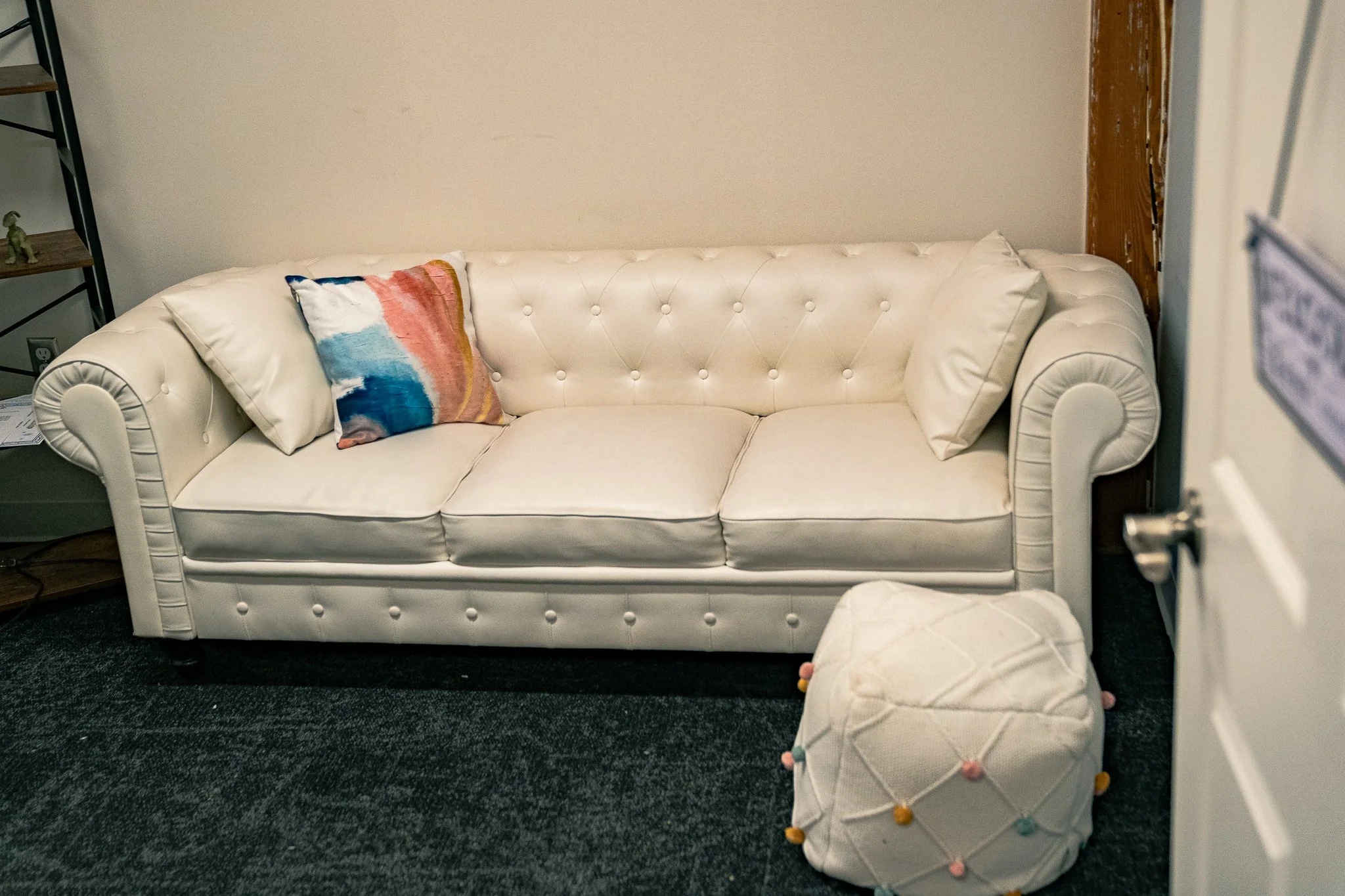 A white tufted sofa with two large pillows and a colorful throw pillow with a painted pattern. There is a small round ottoman with colorful pom-pom trim in front of the sofa, and a corner of a bookshelf with a decorative item on it to the left.
