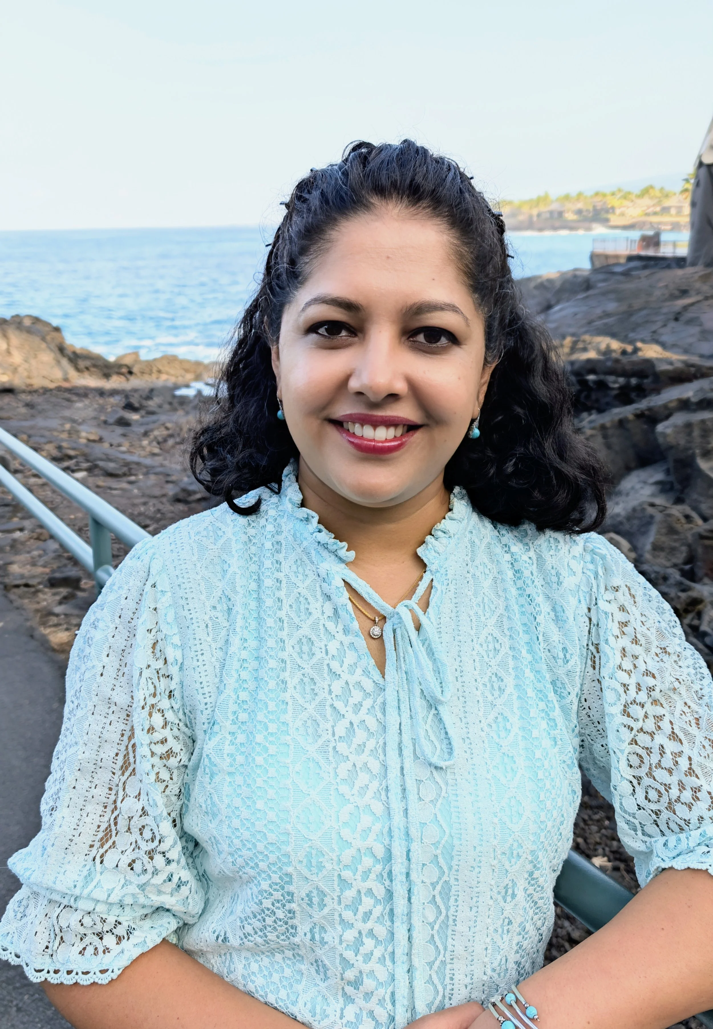 A woman with black curly hair smiling at the camera, wearing a light blue lace blouse, earrings, and jewelry, standing near a rocky shoreline with the ocean and distant land visible in the background.