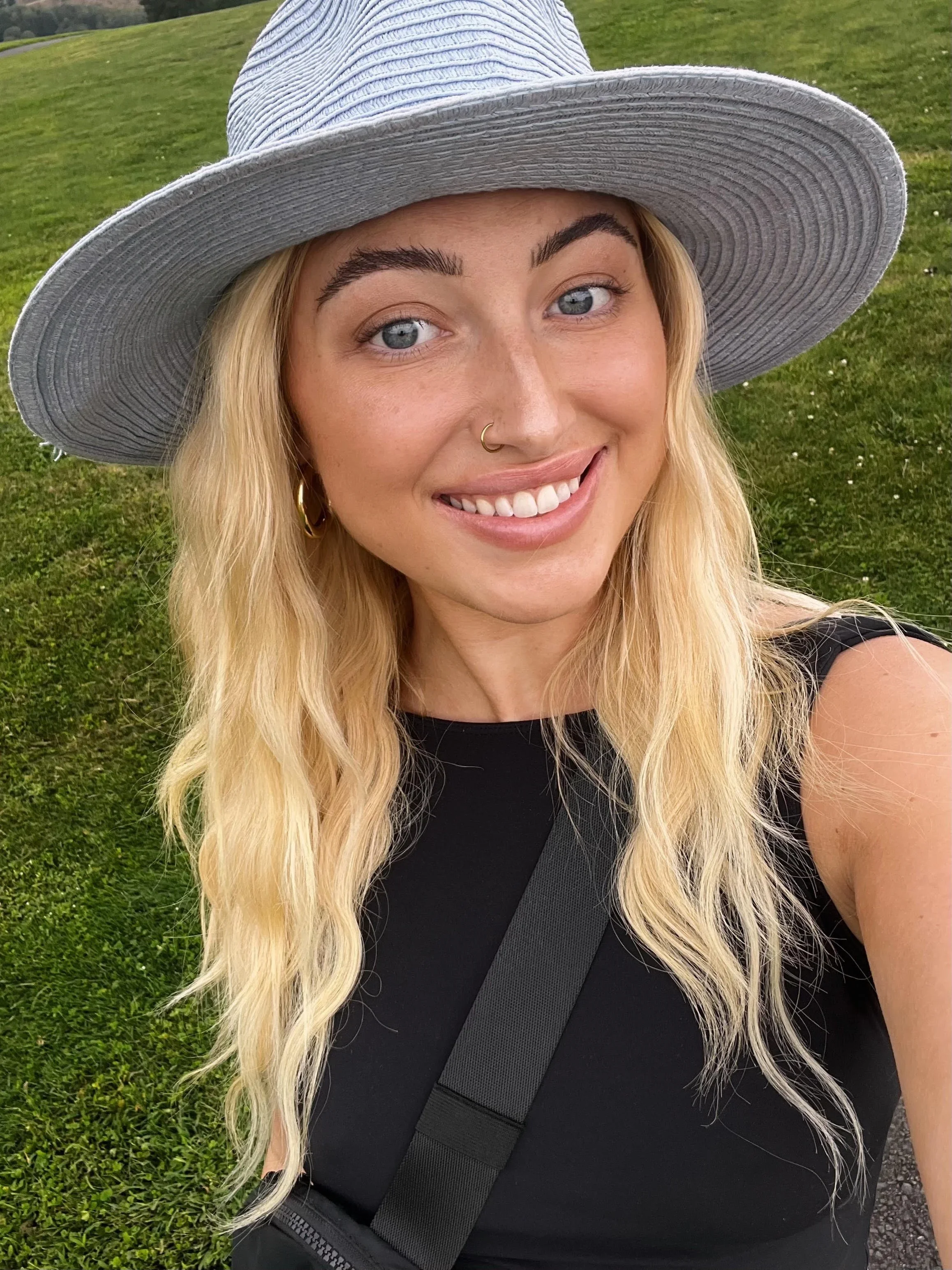 Close-up of a smiling woman with long blonde hair wearing a wide-brimmed gray hat and a black top, standing outdoors on green grass.