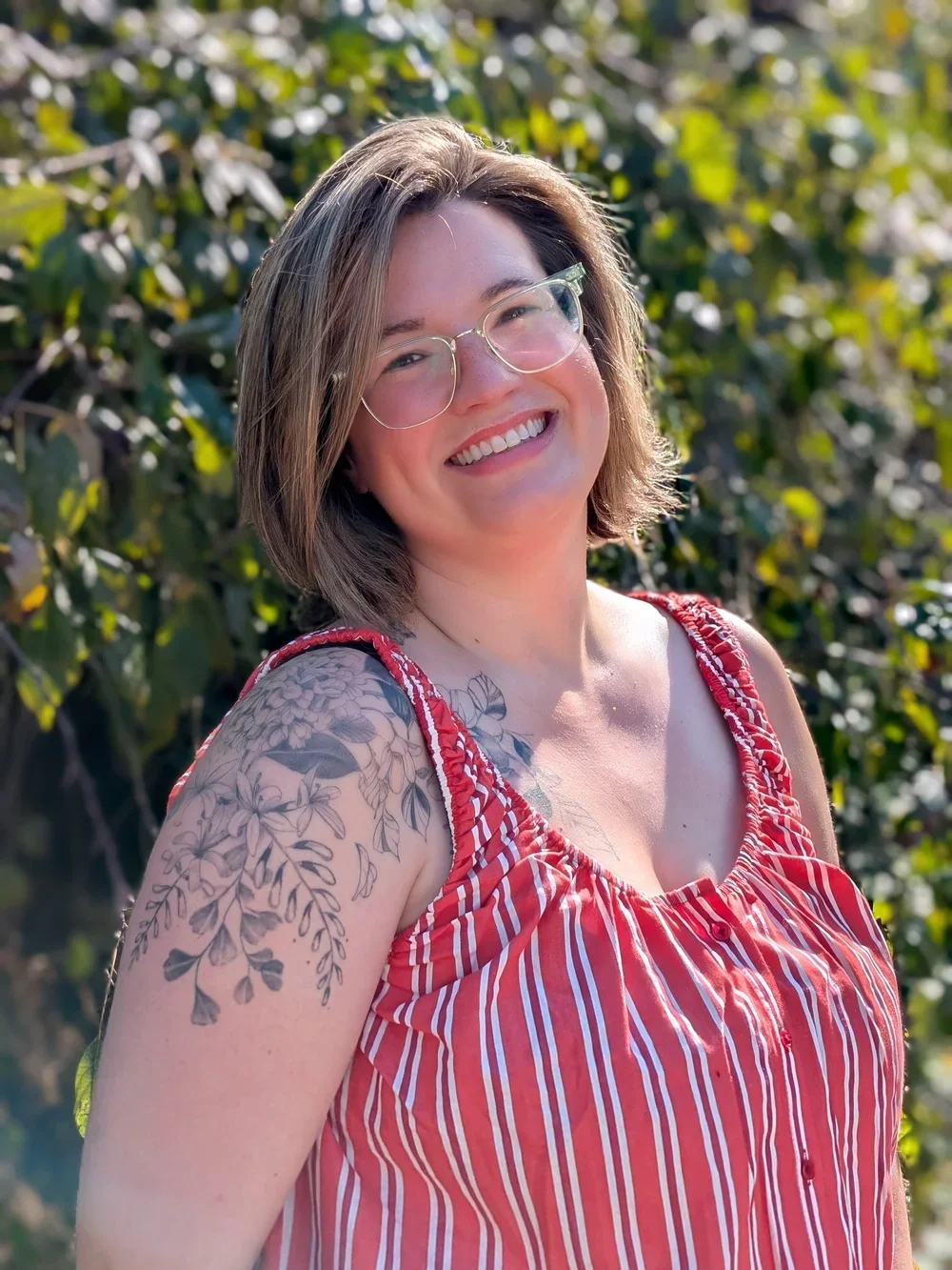 A smiling woman with glasses and short brown hair stands outdoors in front of green foliage, wearing a red and white striped sleeveless top and displaying a floral tattoo on her left shoulder.