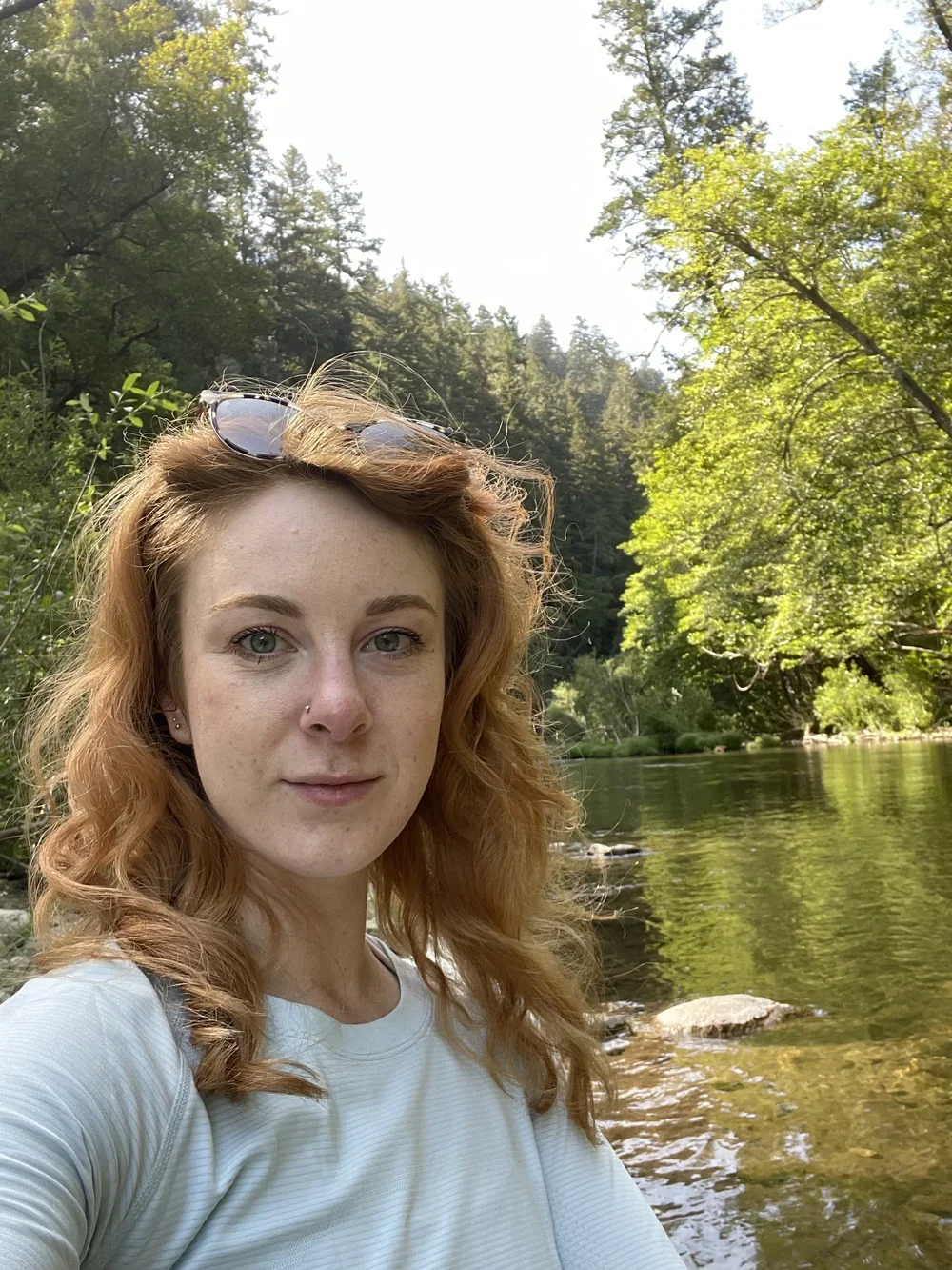 A woman with red wavy hair and a nose piercing taking a selfie by a river surrounded by lush green trees on a sunny day.