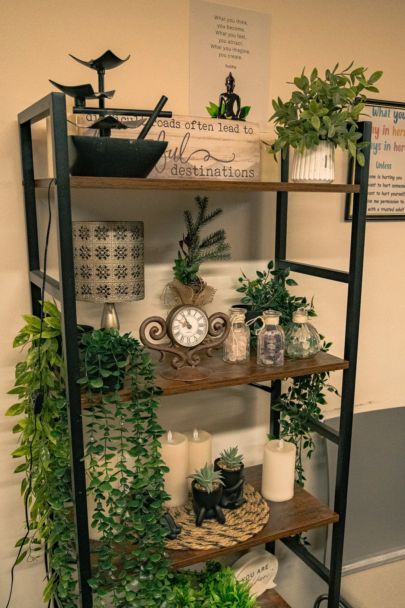 A black metal and wood shelf decorated with plants, candles, a clock, a lamp, and decorative items, with motivational posters on the wall behind.