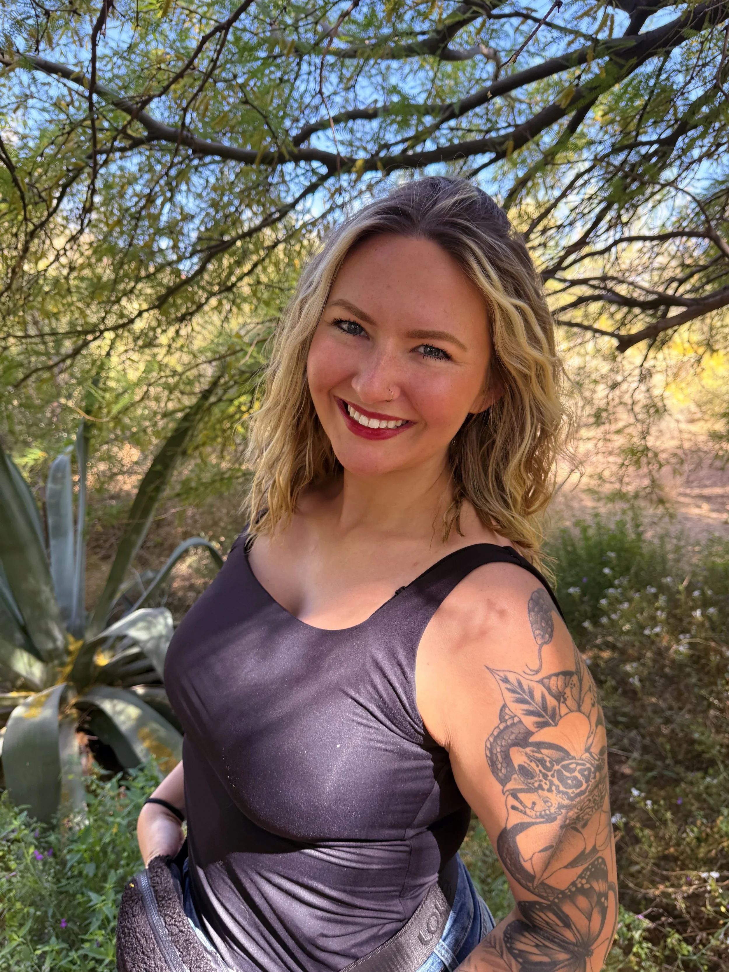 A smiling woman with blonde curly hair, wearing a black sleeveless top, standing outdoors among trees and greenery.