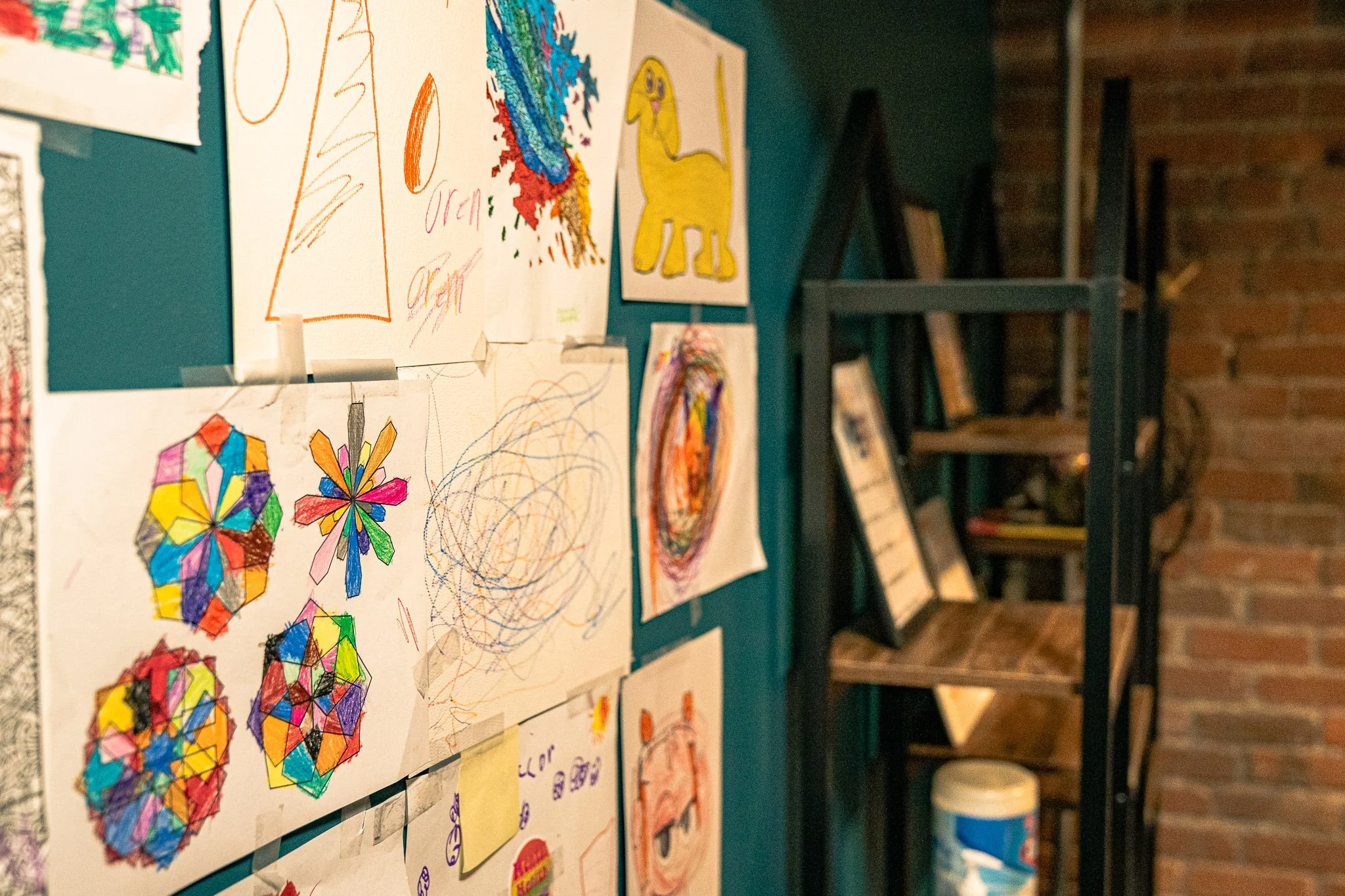 Colorful children's drawings and scribbles taped to a teal wall in a classroom or art space, with a black metal shelf and brick wall visible in the background.