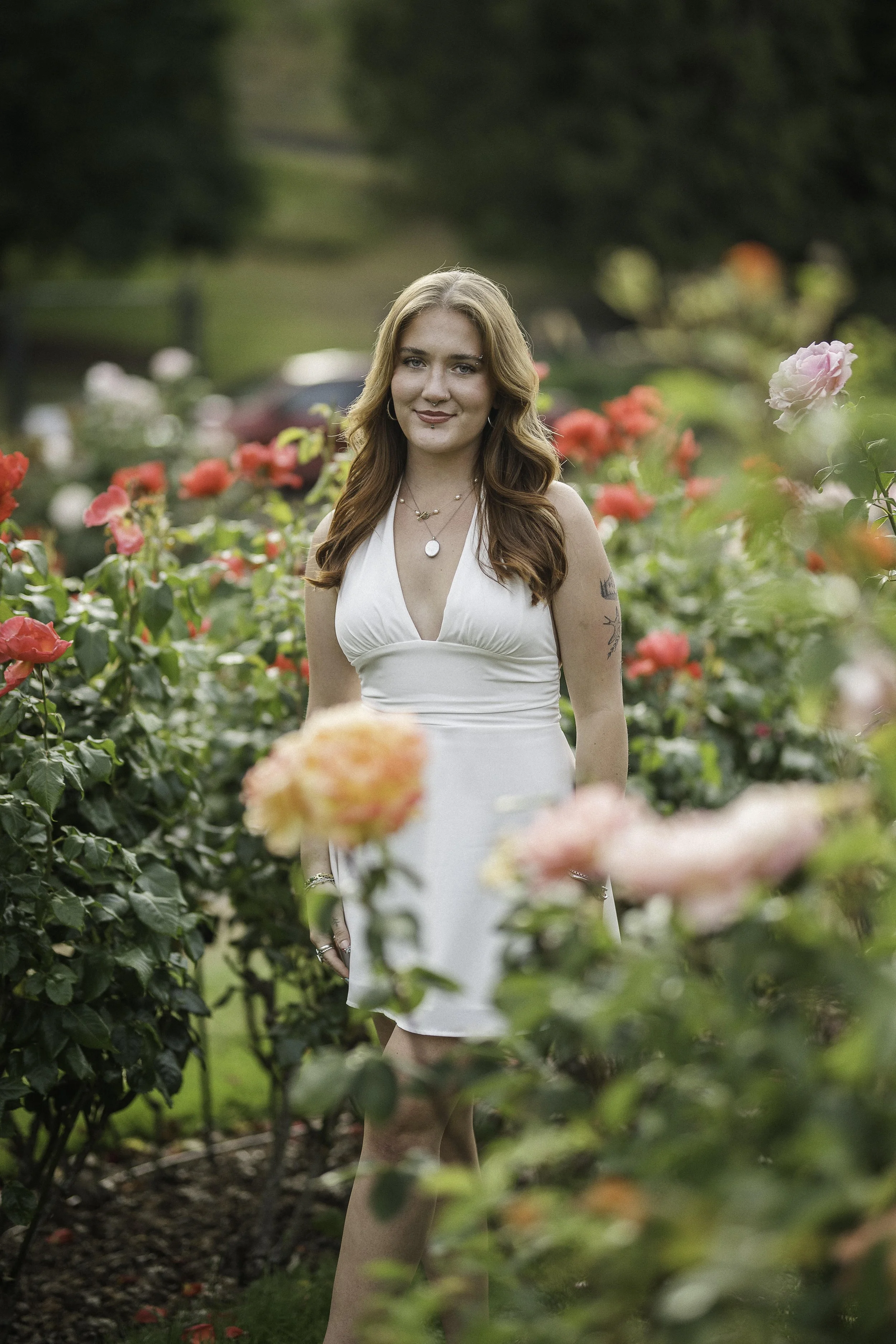 A young woman with long red hair wearing a white dress is standing among pink and red roses in a garden.