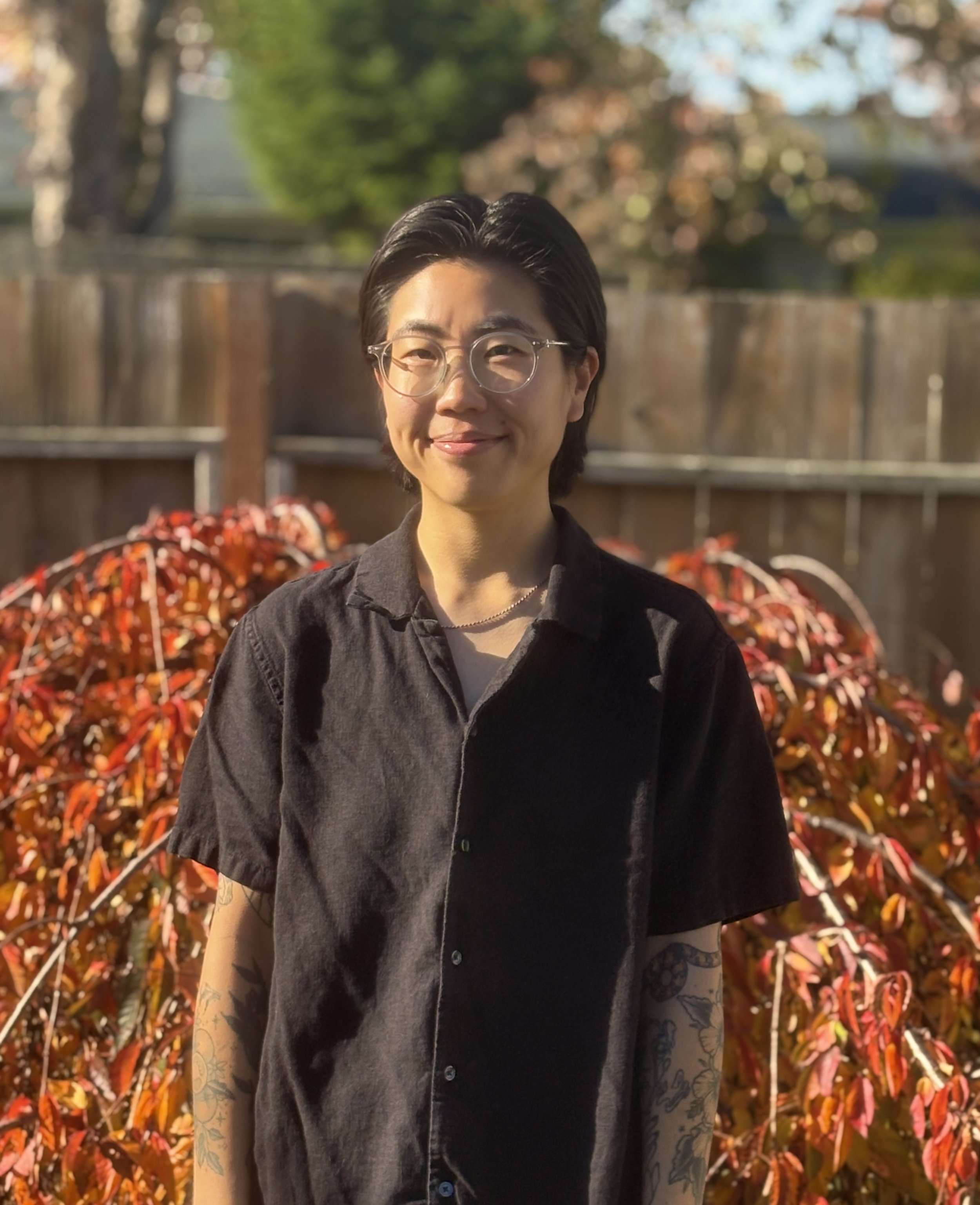 A person with short dark hair, glasses, and tattoos on their arms, standing outdoors in front of colorful autumn leaves, smiling slightly.